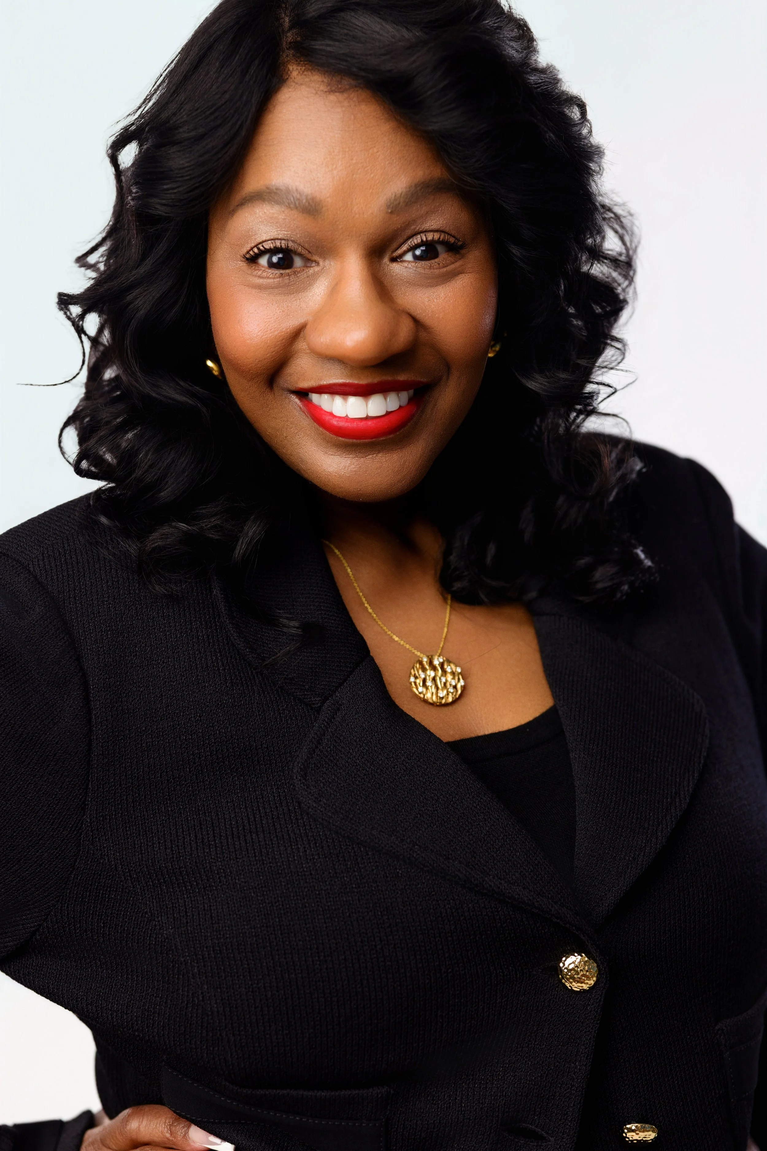 A woman with dark curly hair and dark skin, smiling, wearing red lipstick, gold earrings, a gold pendant necklace, and a black coat with gold buttons, against a plain light background.