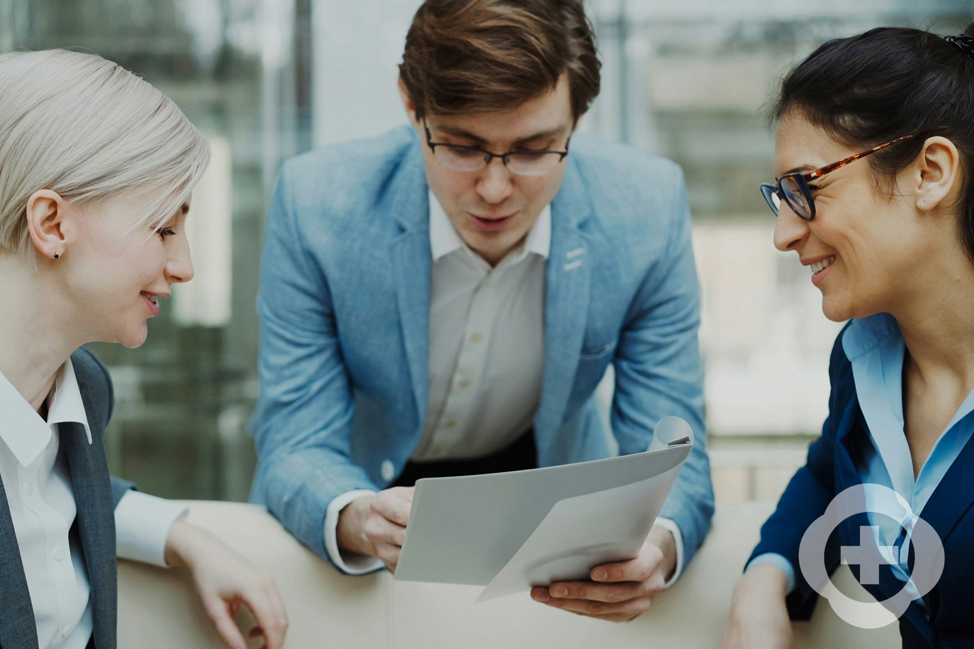 Three business professionals, two women and one man, are engaged in a discussion looking at a document or tablet, seated at a conference table in a bright office.