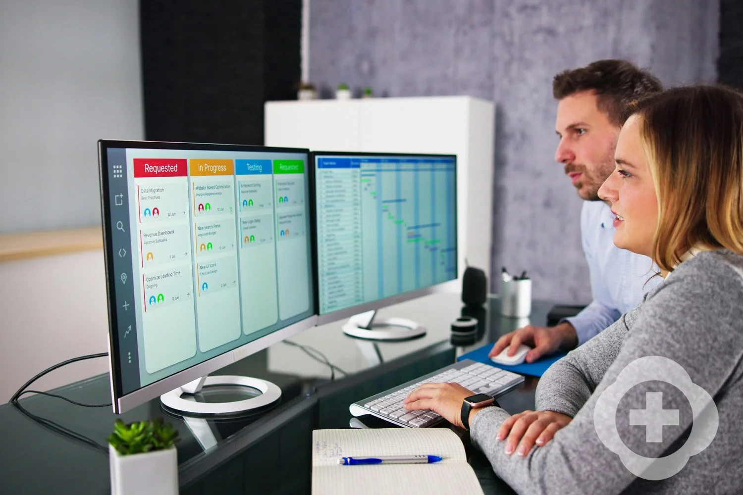 Two people working at desks with dual computer monitors showing project management and scheduling software. One person is using a keyboard and mouse, a notebook and pen are nearby, and the workspace has a small potted plant and office supplies.