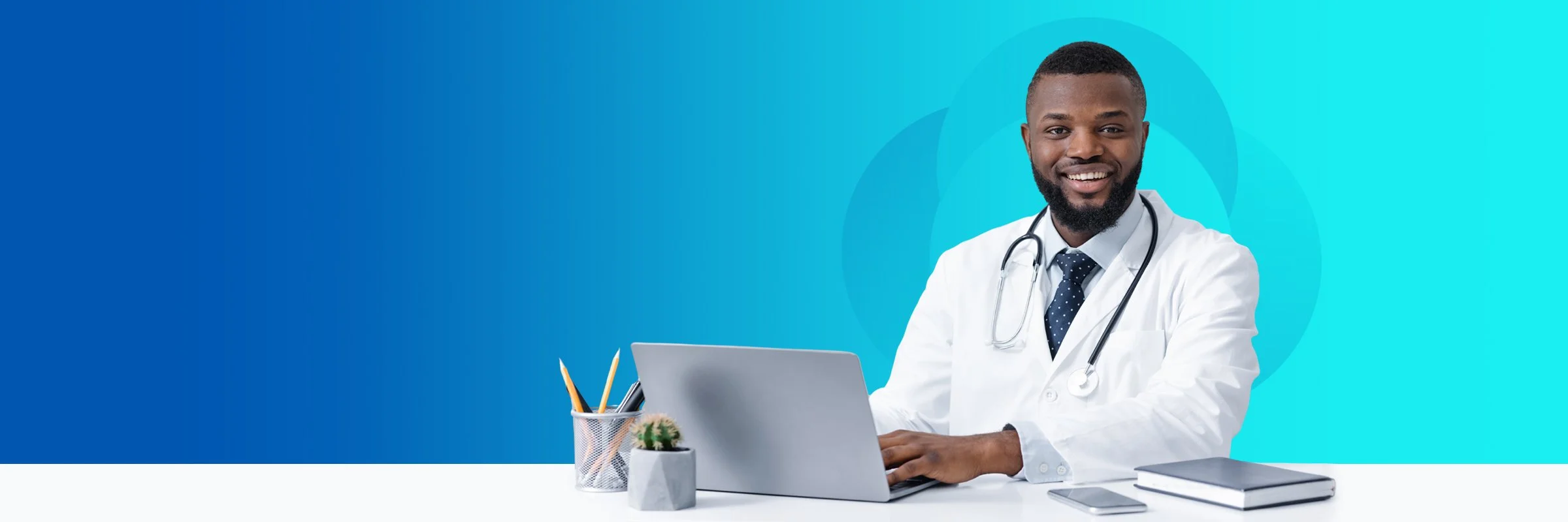 Smiling male doctor with a stethoscope around his neck sitting at a desk with a laptop, notebook, and small potted plant against a blue background.