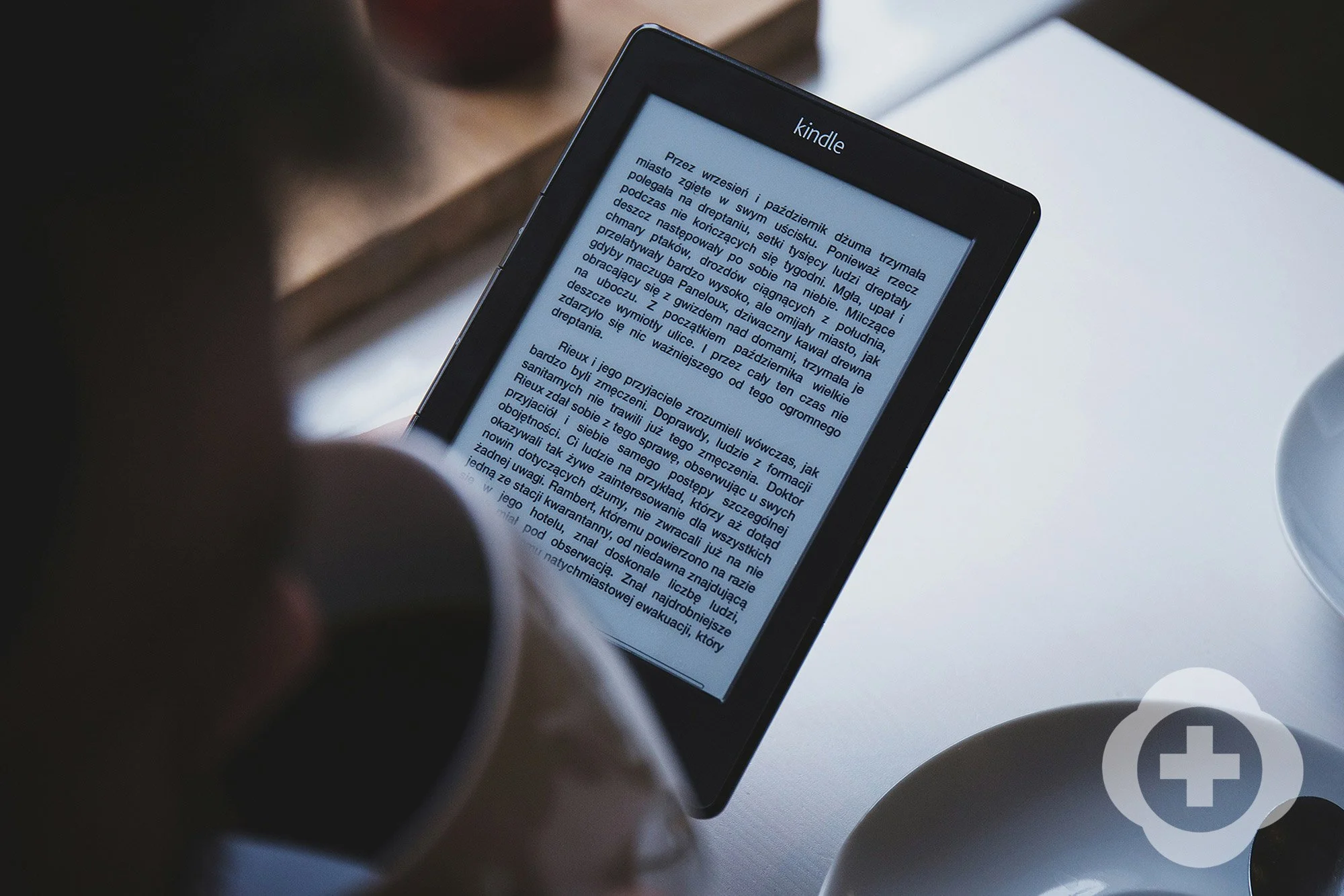 An e-reader device displaying a page of text in Polish, held by a person's hand, with a desk surface and a partial coffee mug visible in the background.