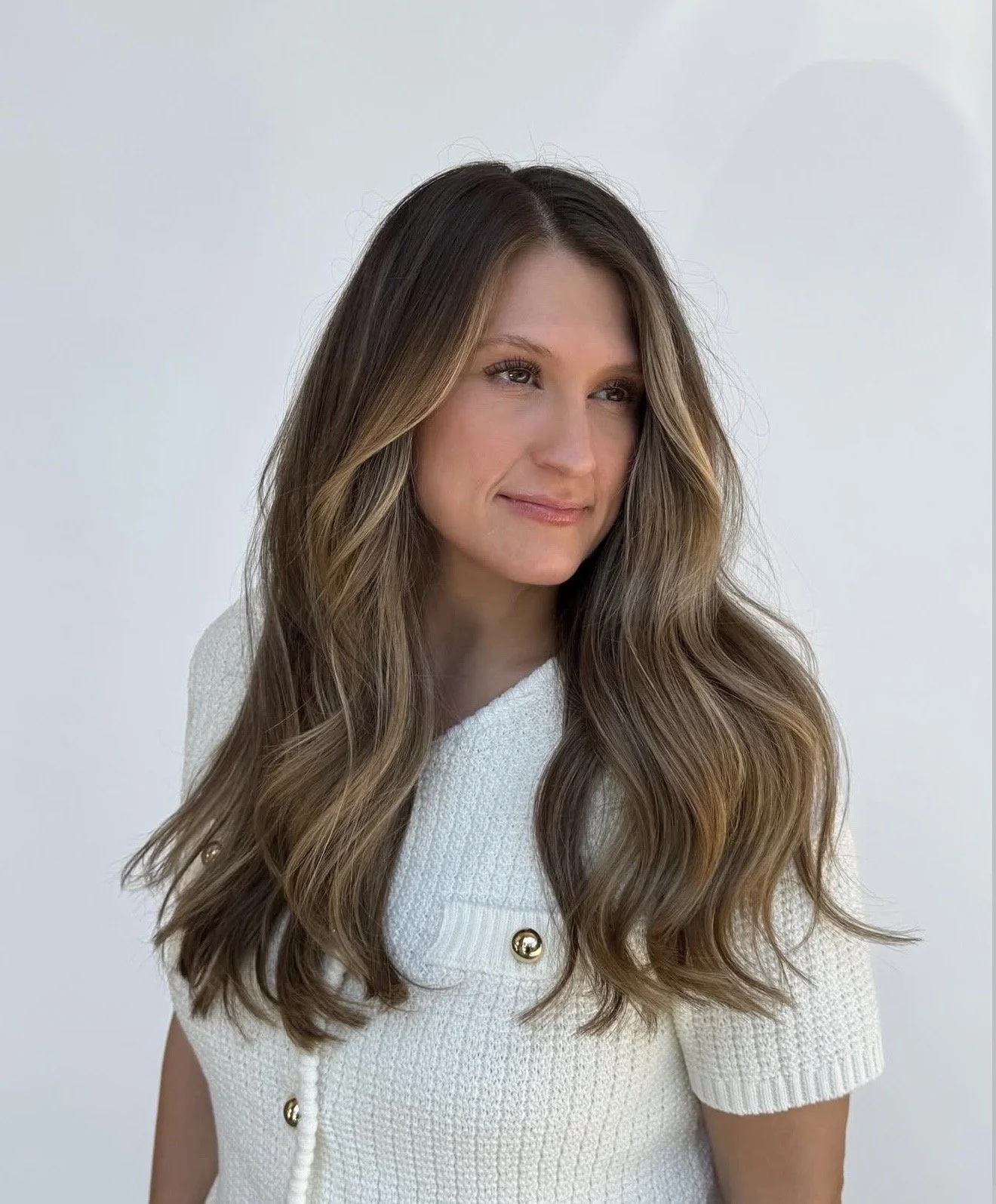 A woman with long, wavy brown hair with blonde highlights, wearing a white textured top with gold buttons, standing against a plain light background.