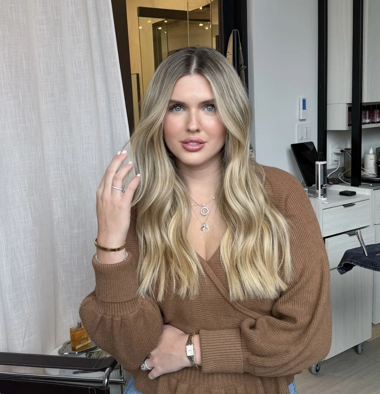 A woman with long blonde wavy hair wearing a brown sweater and jewelry, posing indoors with her hand near her face, in front of a white curtain and a modern interior background.