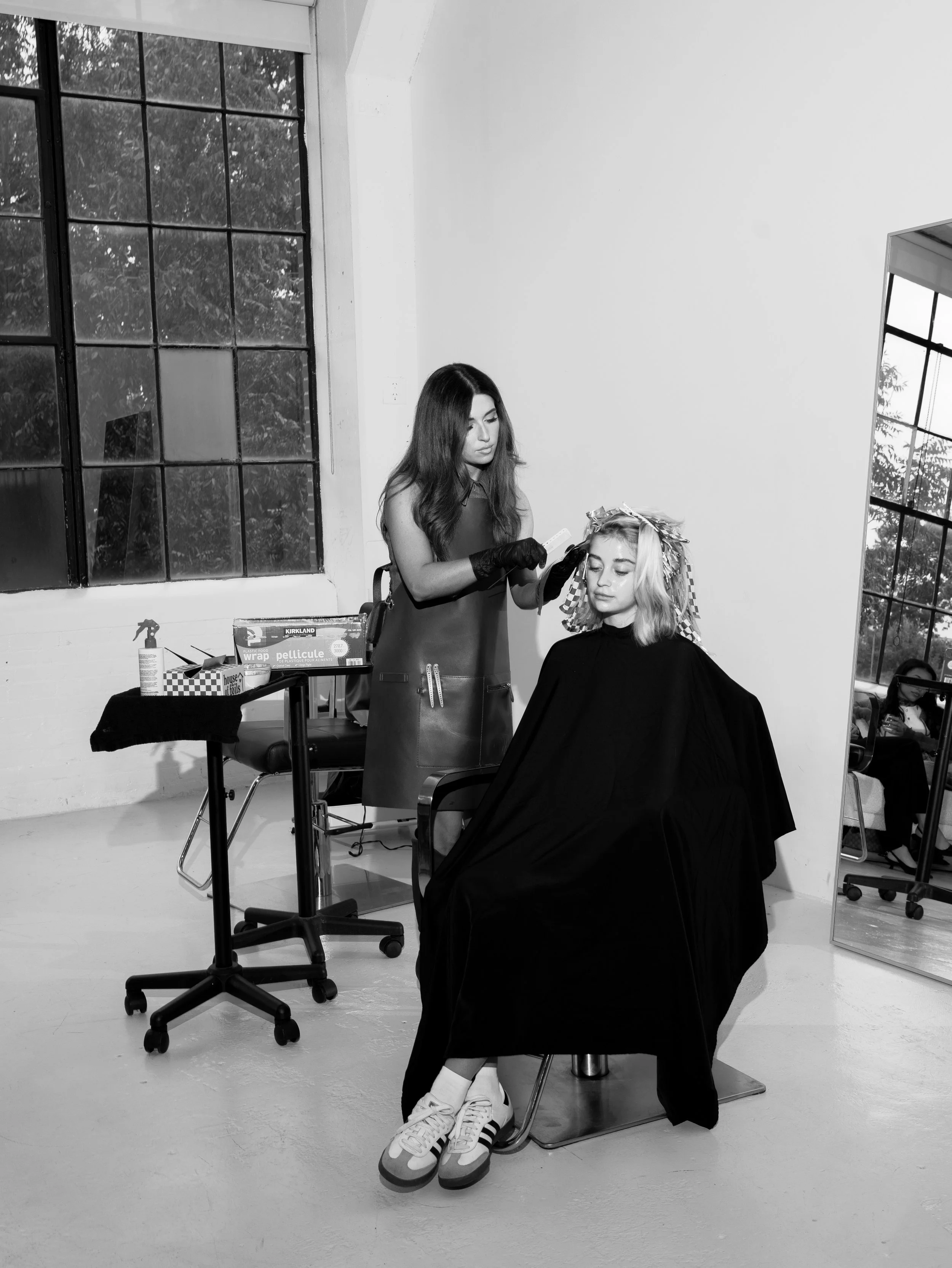 A woman sitting in a salon chair getting her hair styled by a stylist. The stylist is holding a hairbrush and a bottle of spray. The salon has large windows and a mirror reflecting part of the scene.