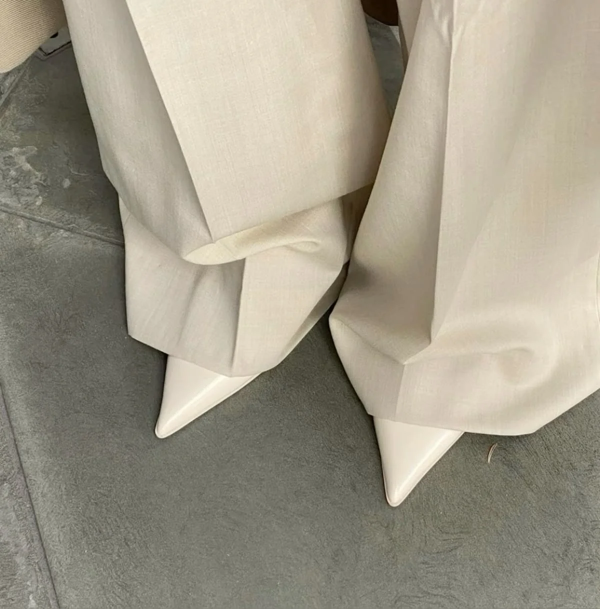 Close-up of the bottom of beige fabric draped over pointed white shoe tips on a gray tiled floor.