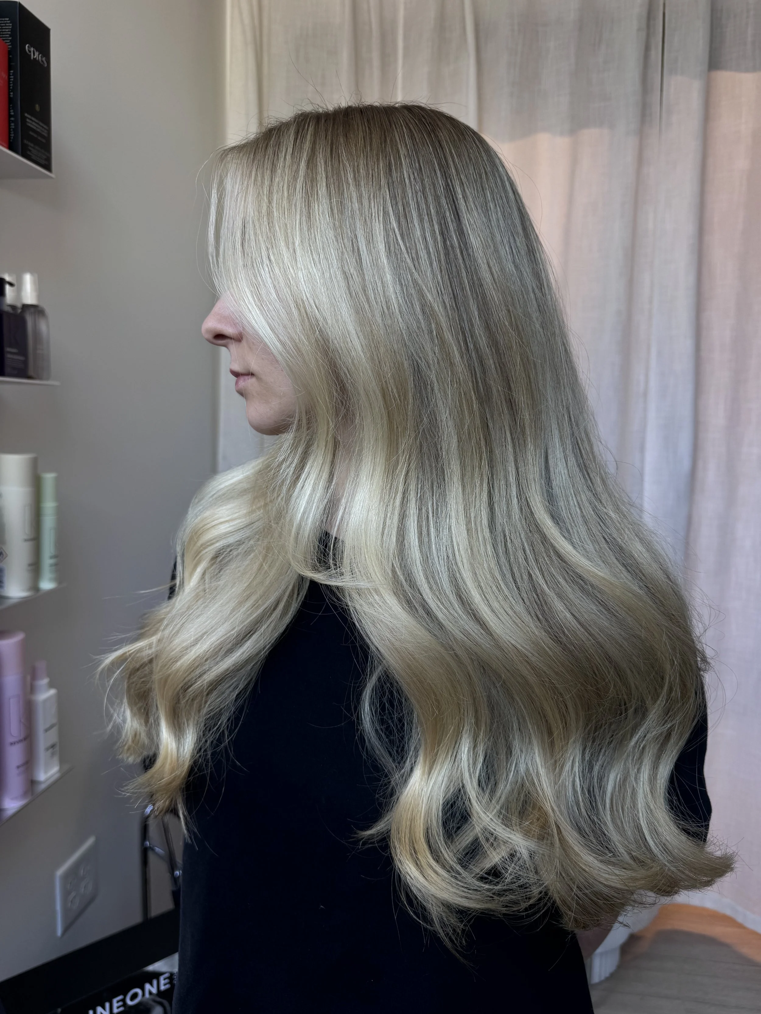 A woman with long, wavy, blonde hair standing in a room with beige curtains and a shelf of hair products.