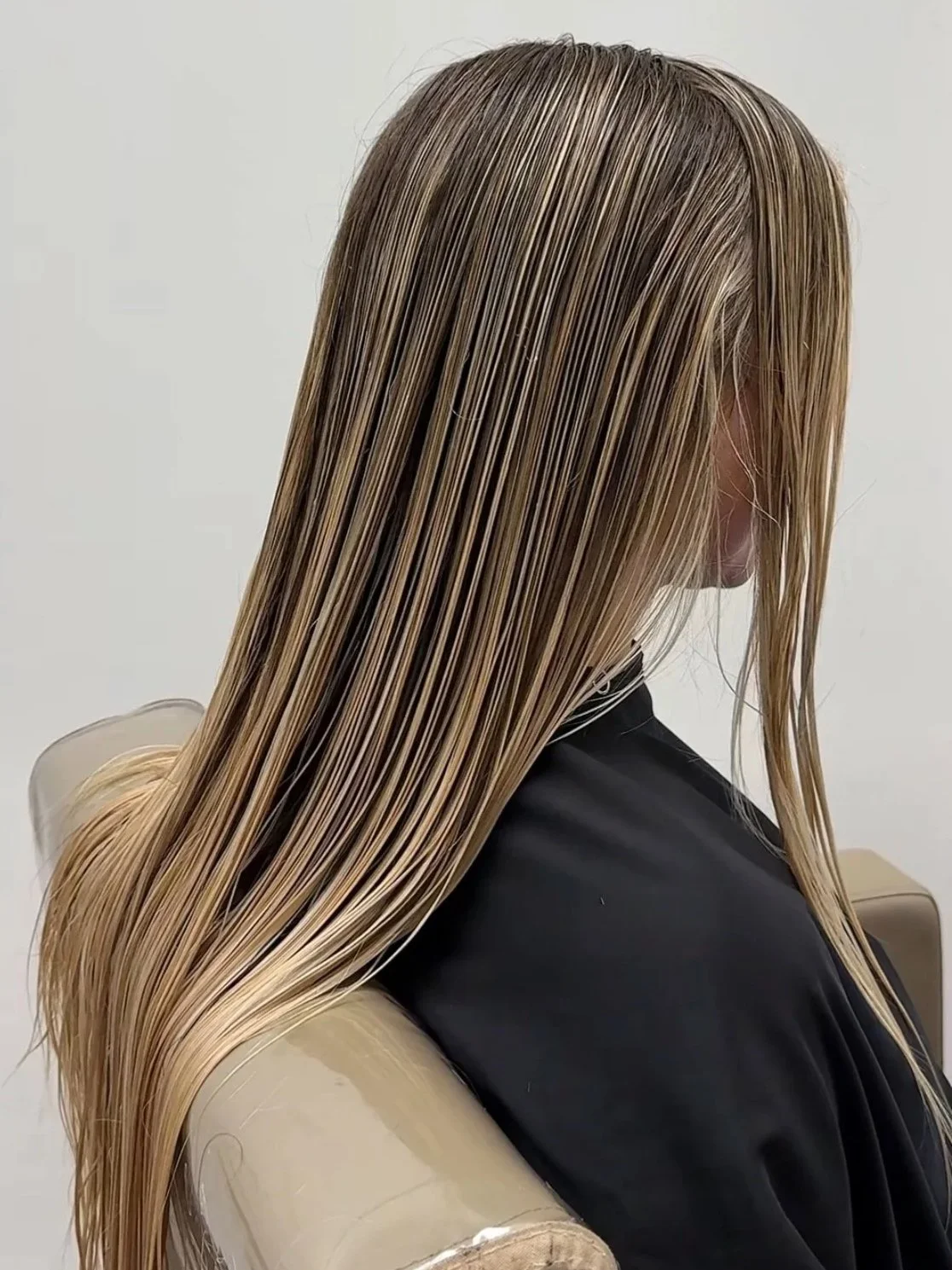 A person with long, straight blonde hair sitting on a beige chair, with hair appearing wet or styled, wearing a black shirt.