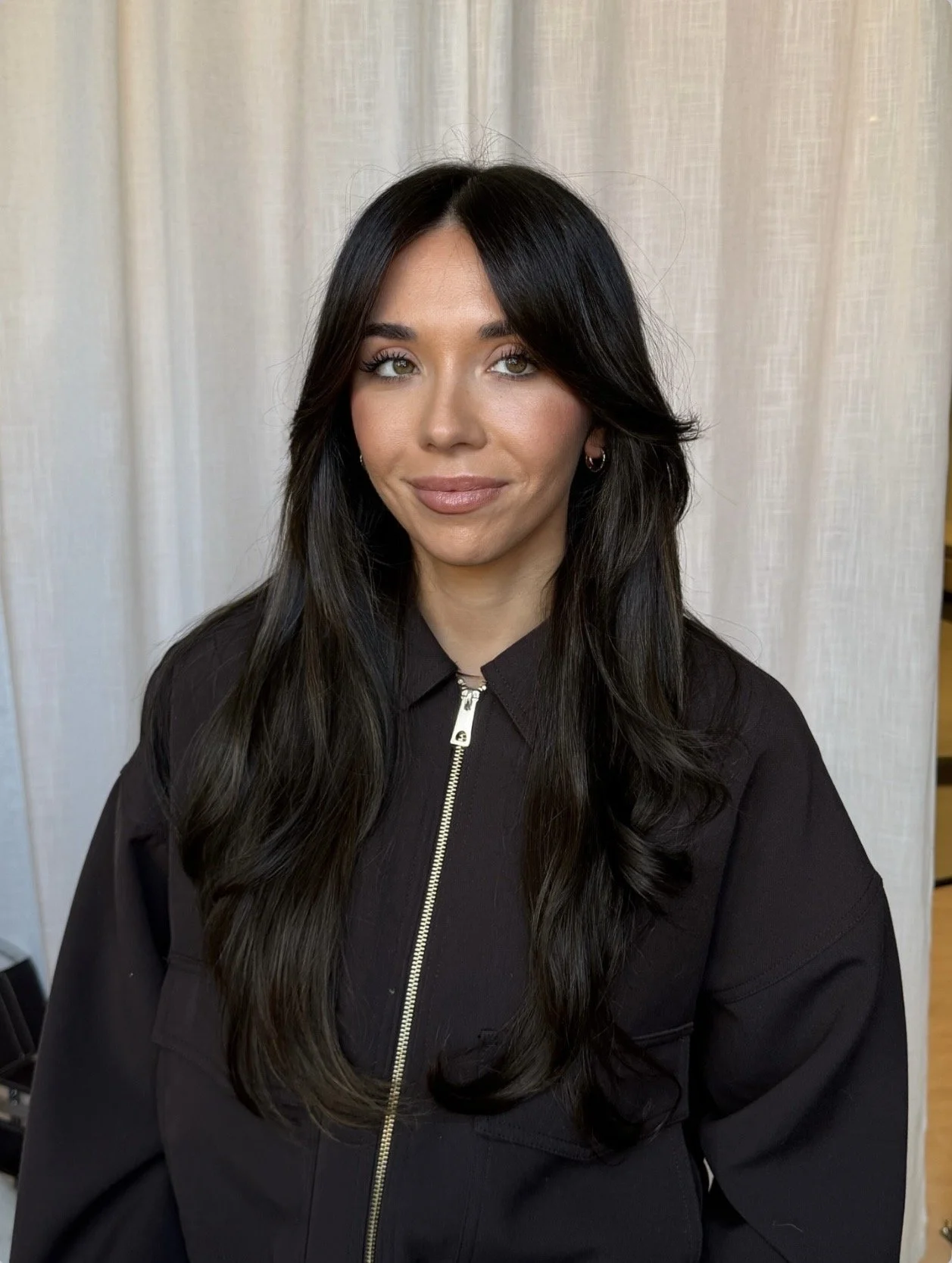 Portrait of a woman with long dark hair, wearing a black zip-up jacket, standing against a light-colored curtain background.