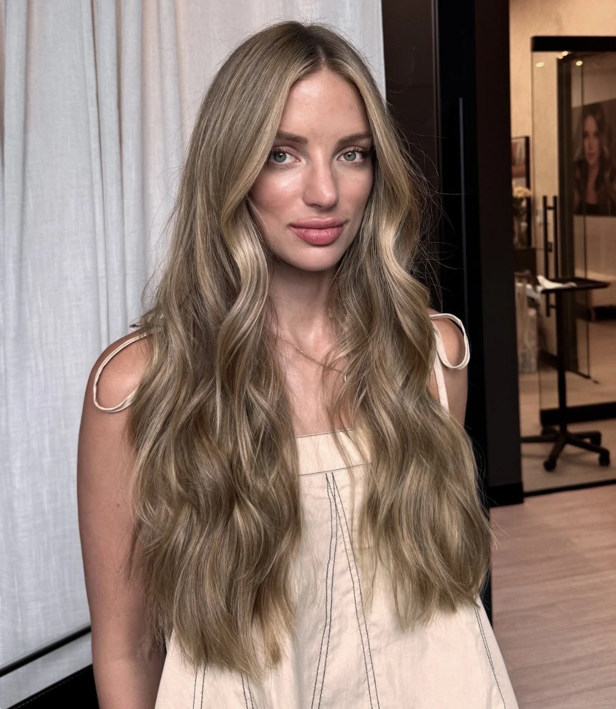 A woman with long, wavy blonde hair, wearing a light-colored dress with tied shoulder straps, stands indoors near a white curtain and a black cabinet. She has a slight smile and is looking at the camera.