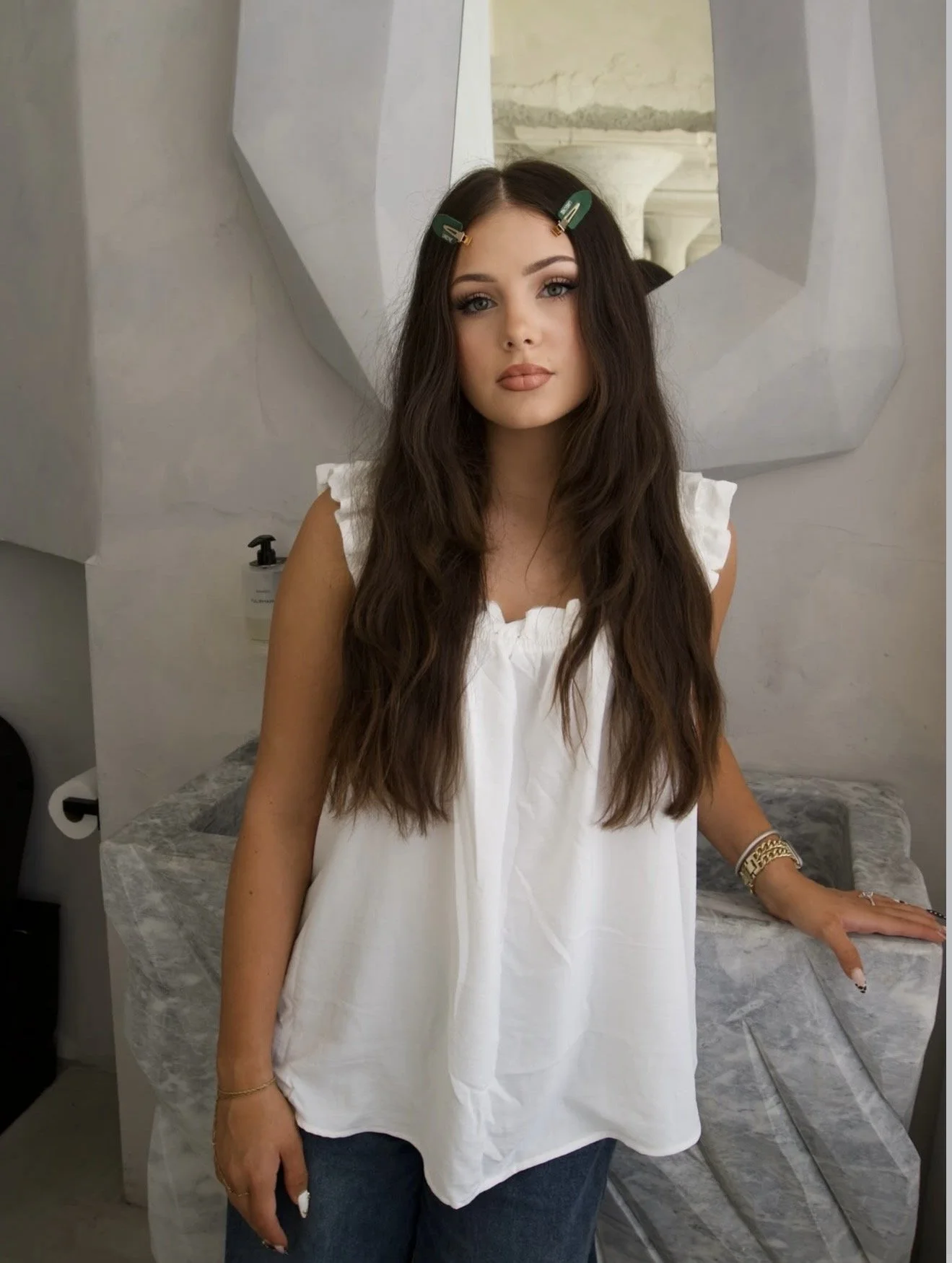 A woman with long wavy brown hair stands in a salon with clips in her hair, wearing a white sleeveless top and blue jeans. She has makeup on and is wearing jewelry, including a bracelet and a ring, leaning against a marble countertop, behind a mirror with decorative arches.