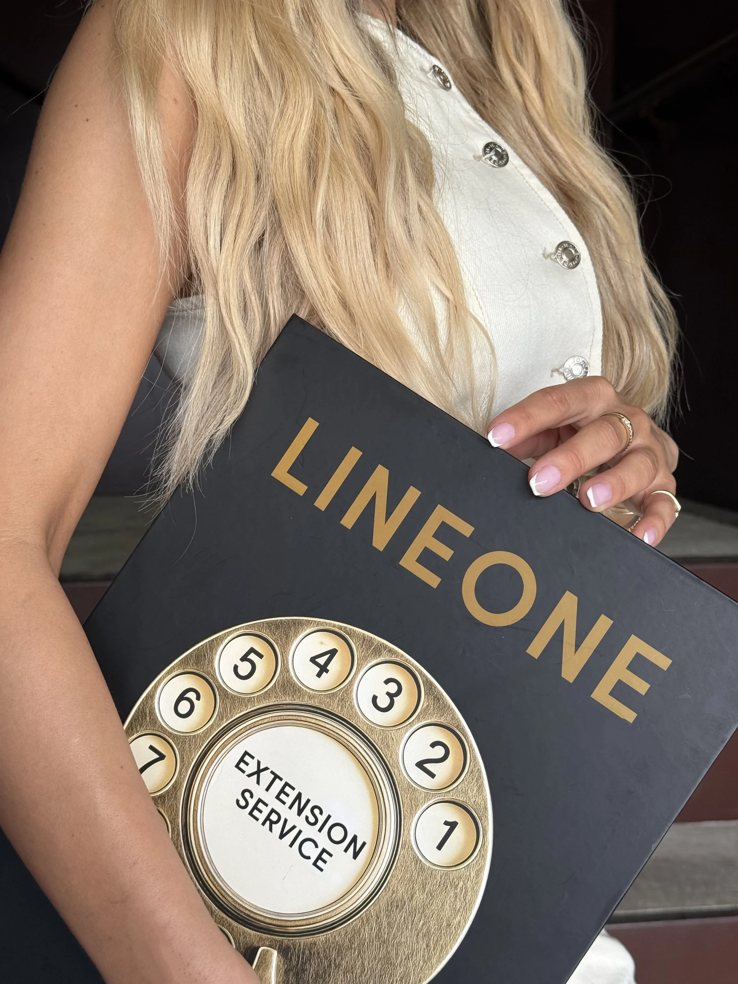 A woman with long blonde hair is holding a black book or folder with the word 'LINEONE' in large gold letters. She is wearing a white shirt with shiny buttons, a ring on her ring finger, and is standing next to a gold-colored lottery or bingo ball with numbers and the words 'Extension Service' on it.