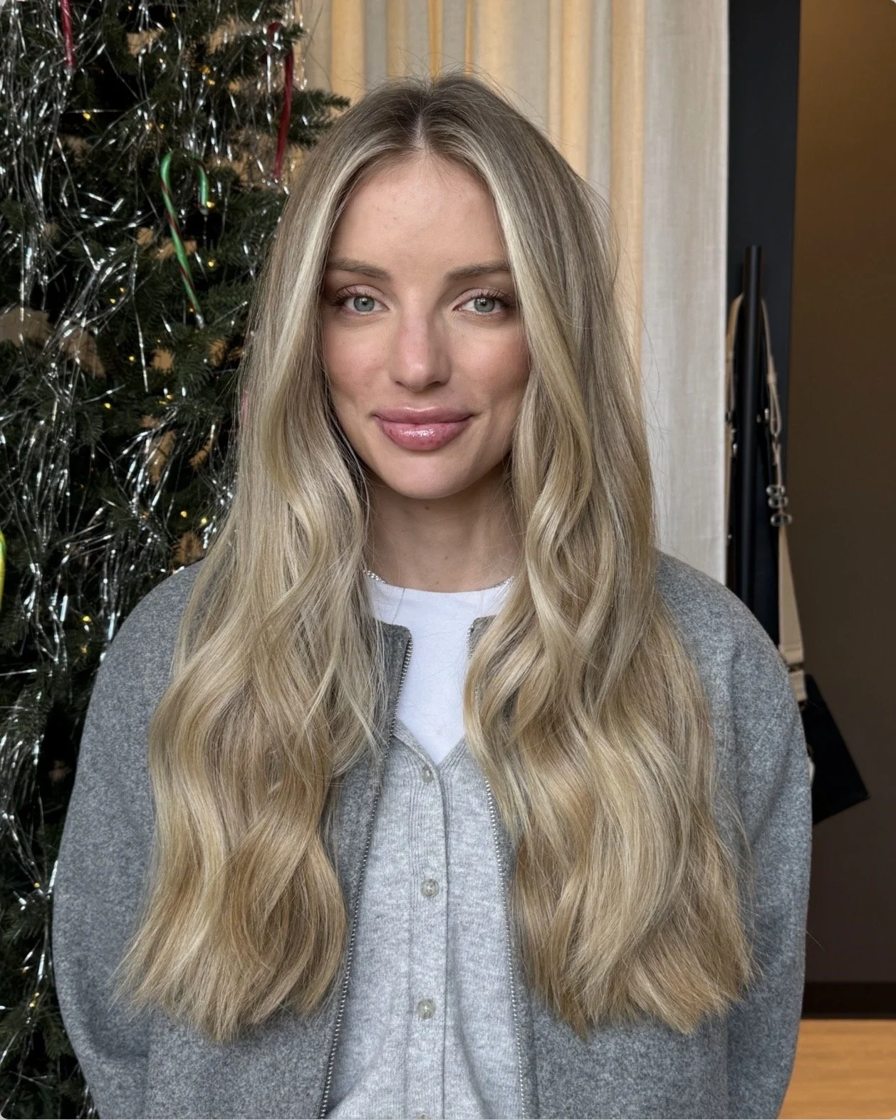 A young woman with long, wavy blonde hair and blue eyes, smiling softly in front of a decorated Christmas tree.