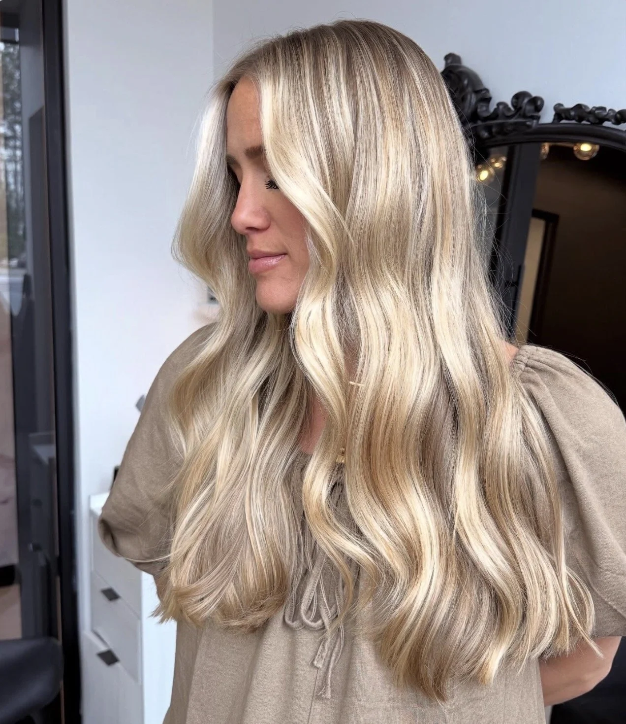 A woman with long, wavy blonde hair, looking down, standing indoors near a black-framed mirror with lights.