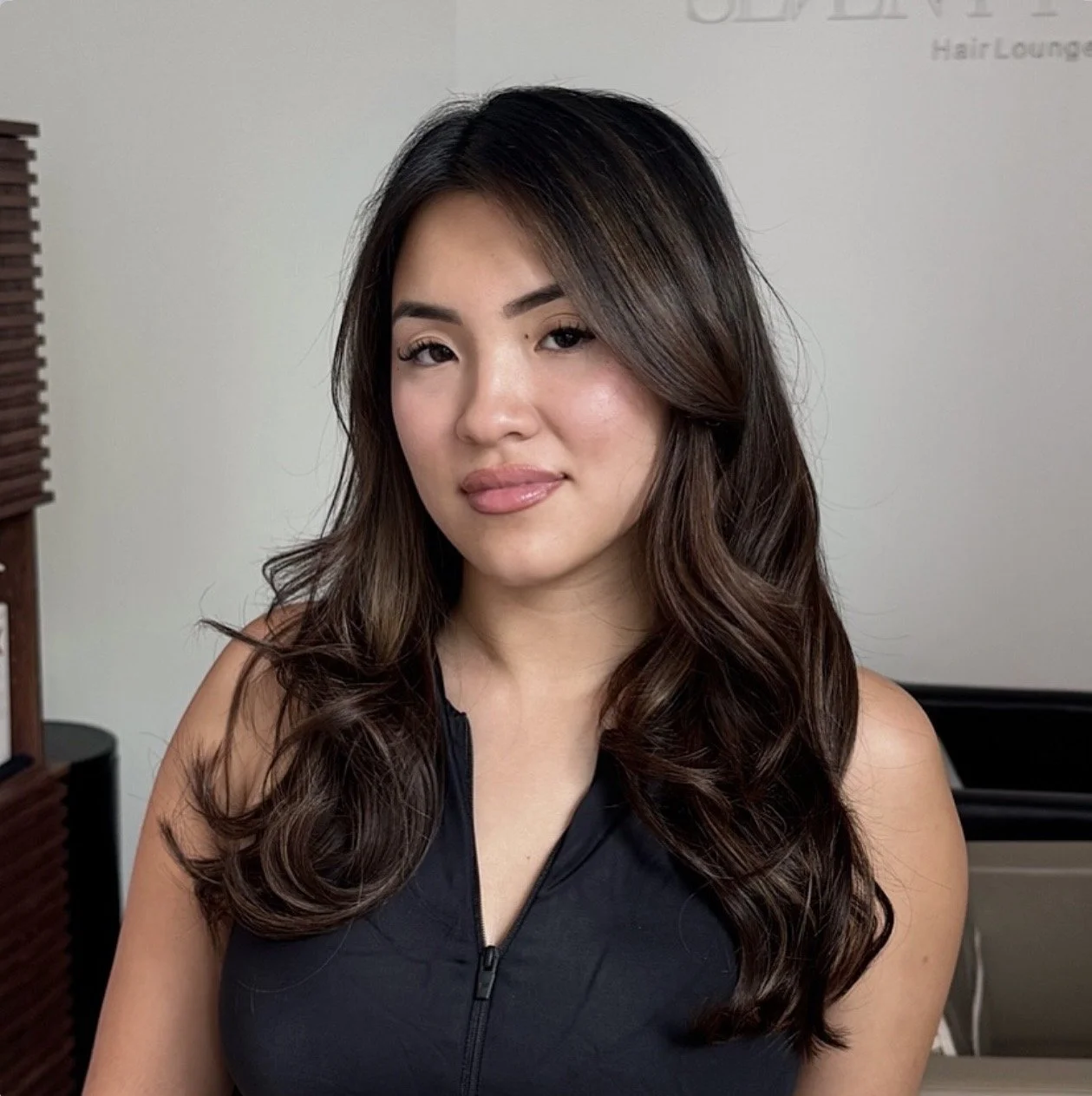 A woman with long wavy brown hair, wearing a sleeveless black top with a zipper, looking at the camera with a slight smile.
