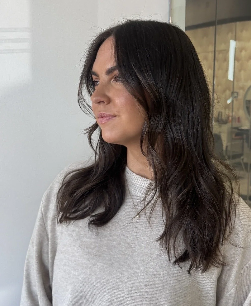 Side view of a woman with long, wavy dark brown hair, wearing a light beige sweater and a delicate necklace, standing indoors with a blurred background.
