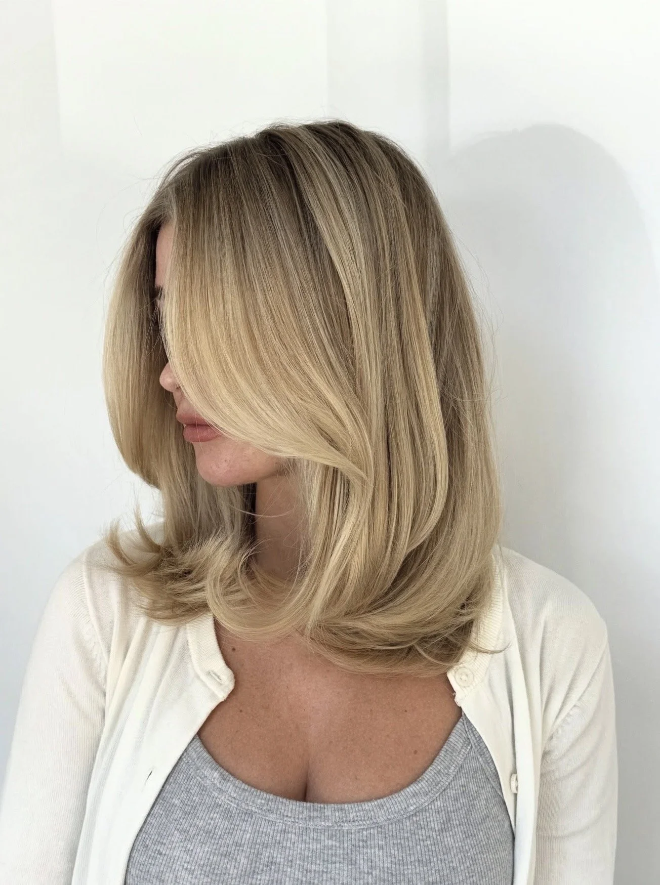 Woman with shoulder-length blonde hair styled in loose waves, wearing a light gray shirt and a white cardigan, standing against a plain white wall.