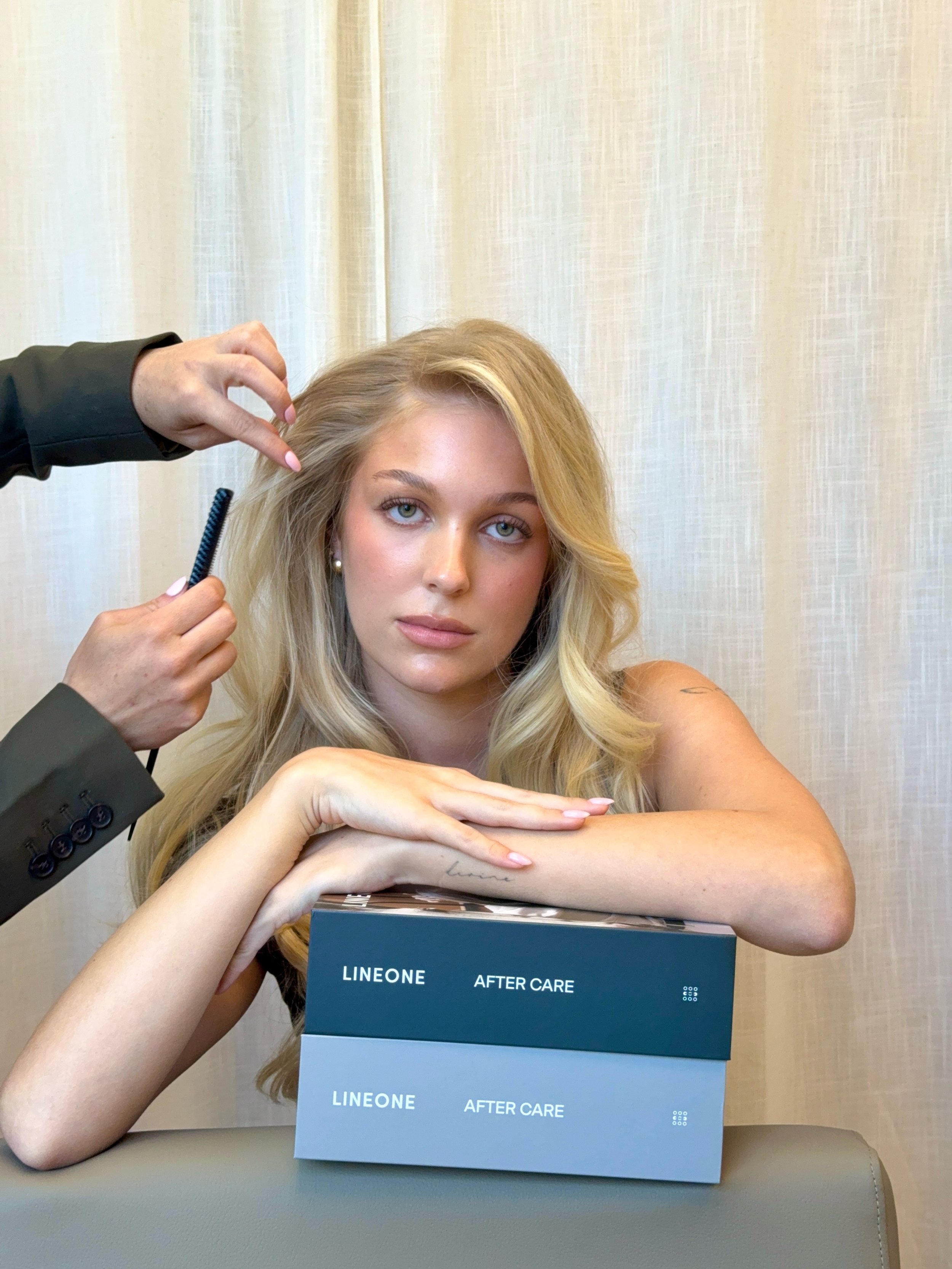 A woman with long blonde hair sitting at a table, resting her arms on a box labeled "LINEONE AFTER CARE." A person, whose face is not visible, stands beside her and is holding a hairbrush and a section of her hair, likely styling or preparing it.