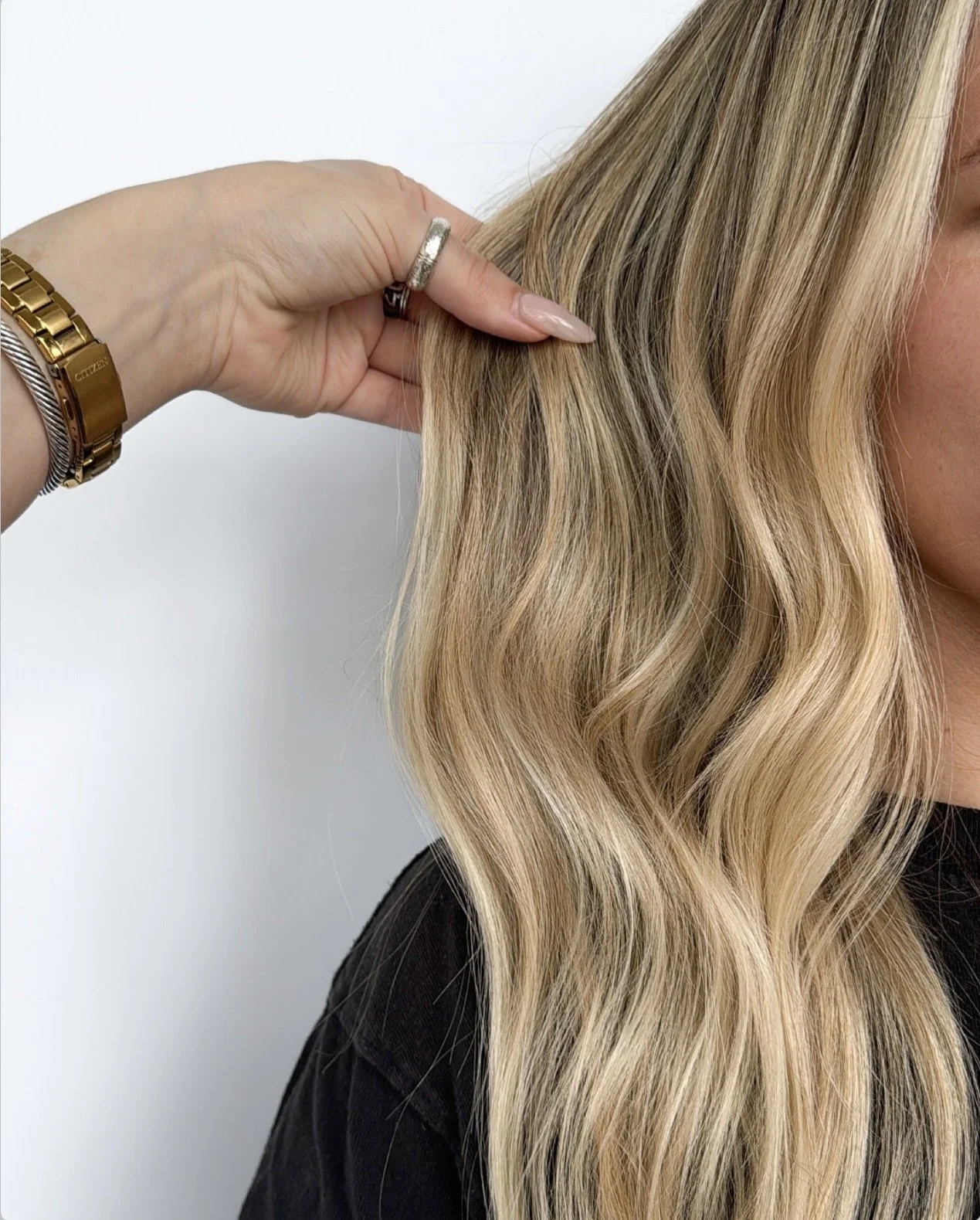 Close-up of a hand touching long, wavy blonde hair with highlights, wearing jewelry including rings and a gold watch, against a plain light background.