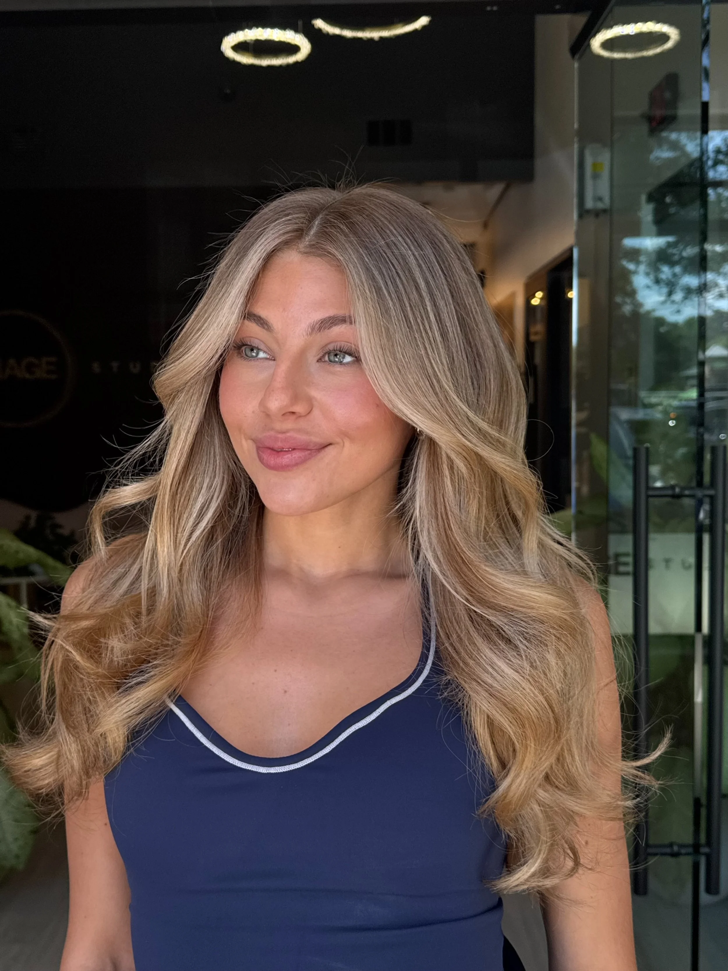 A woman with long, wavy blonde hair, wearing a blue sleeveless top with white piping, standing indoors near a glass door with a reflection of trees outside.