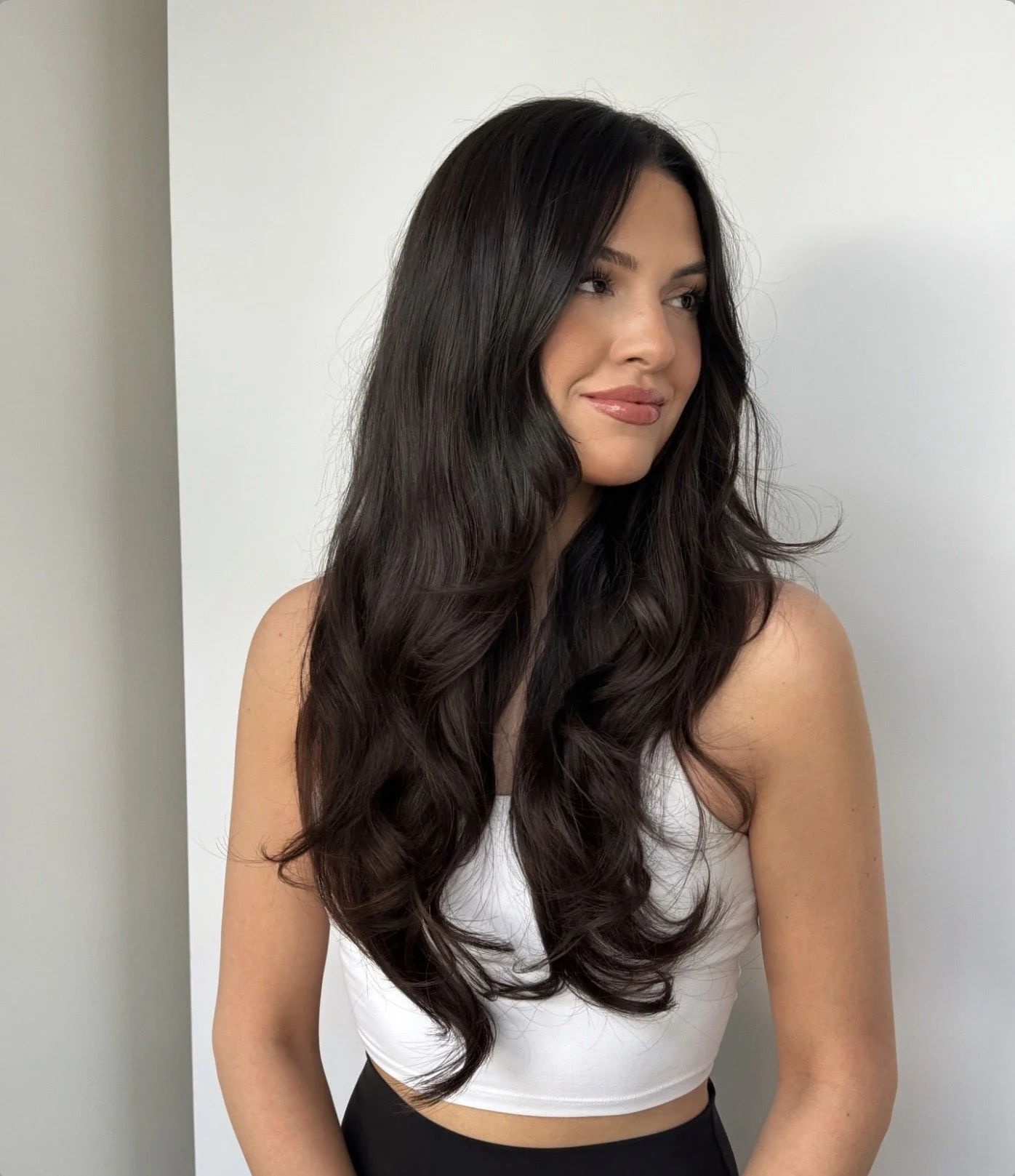 A woman with long, dark, wavy hair wearing a white sleeveless top and black bottom, standing against a plain white background.
