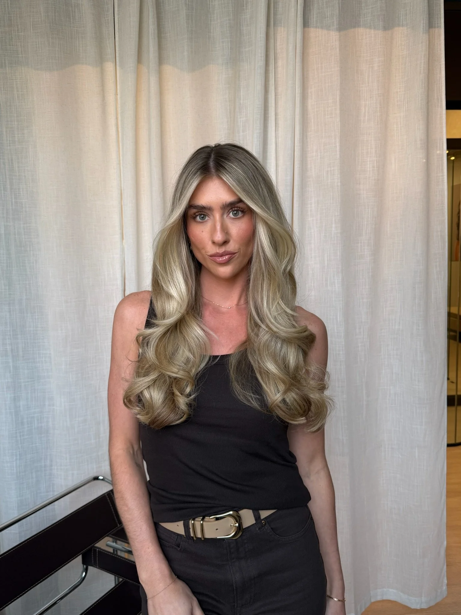 A woman with long, wavy blonde hair, wearing a black tank top and black pants with a beige belt, standing in front of cream-colored curtains.