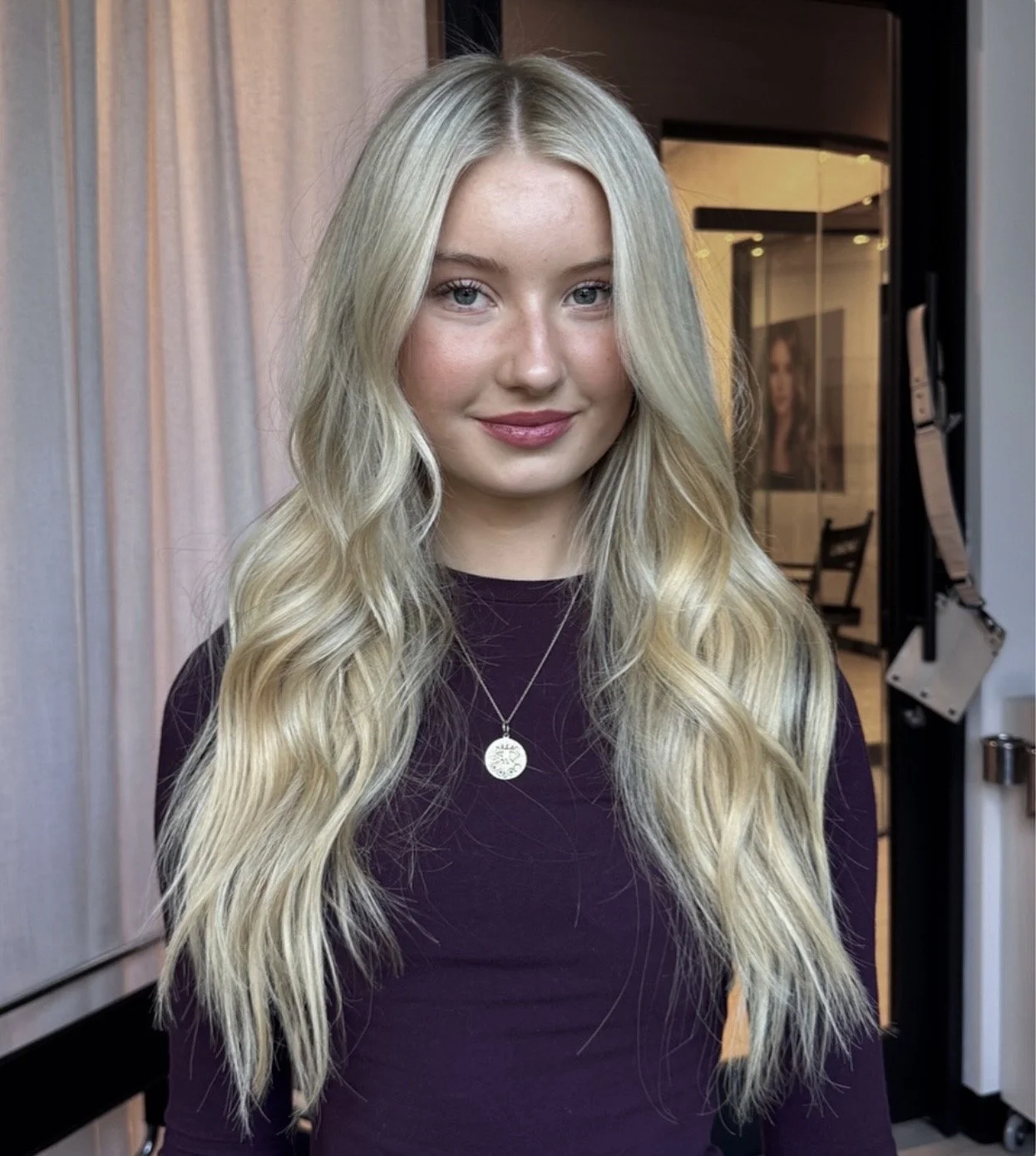 A young woman with long, wavy blonde hair, wearing a dark purple top and a silver necklace, standing indoors near a curtain and a painting of a woman
