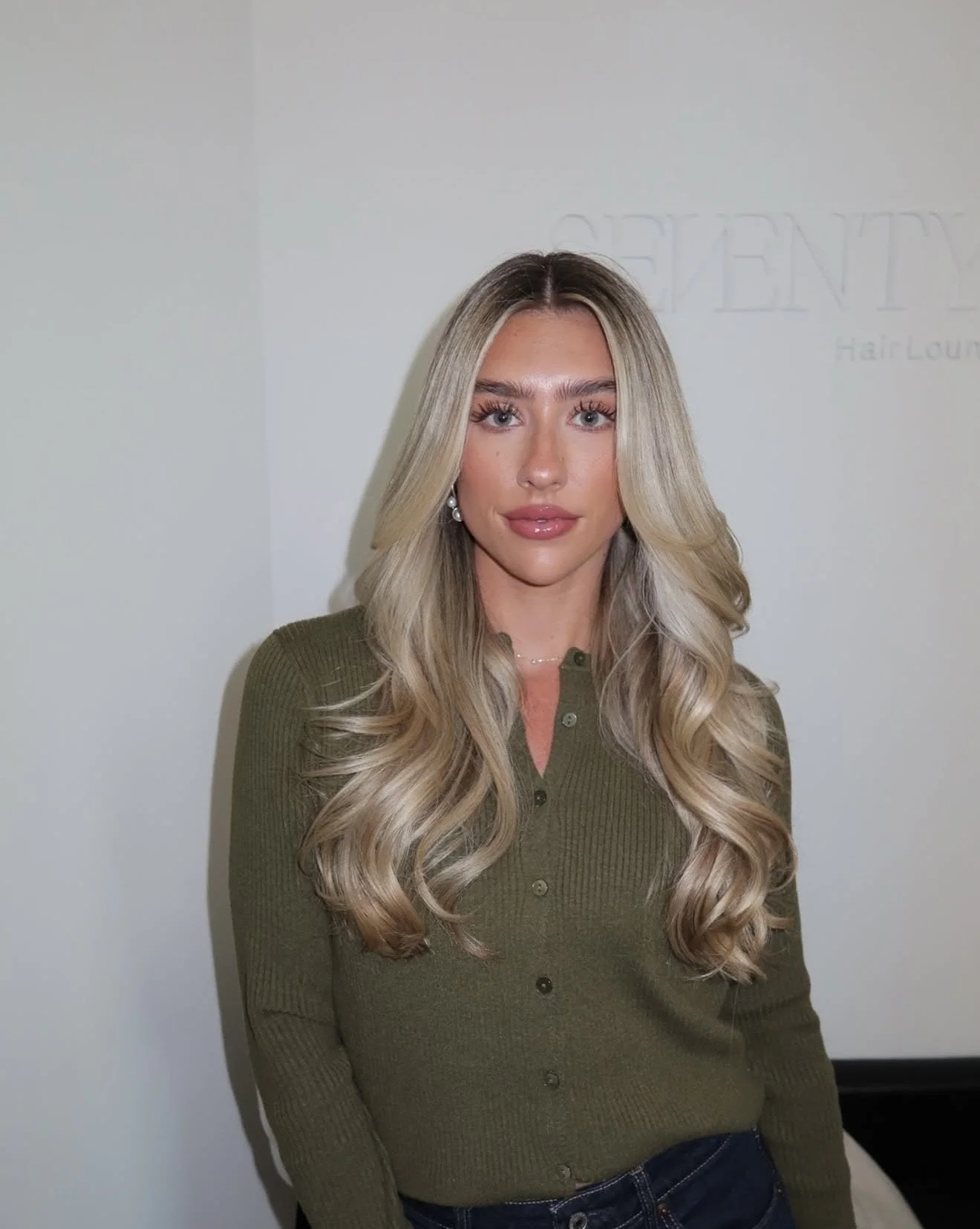 A young woman with long, wavy blonde hair, wearing a green button-up sweater and earrings, standing indoors against a light-colored wall.