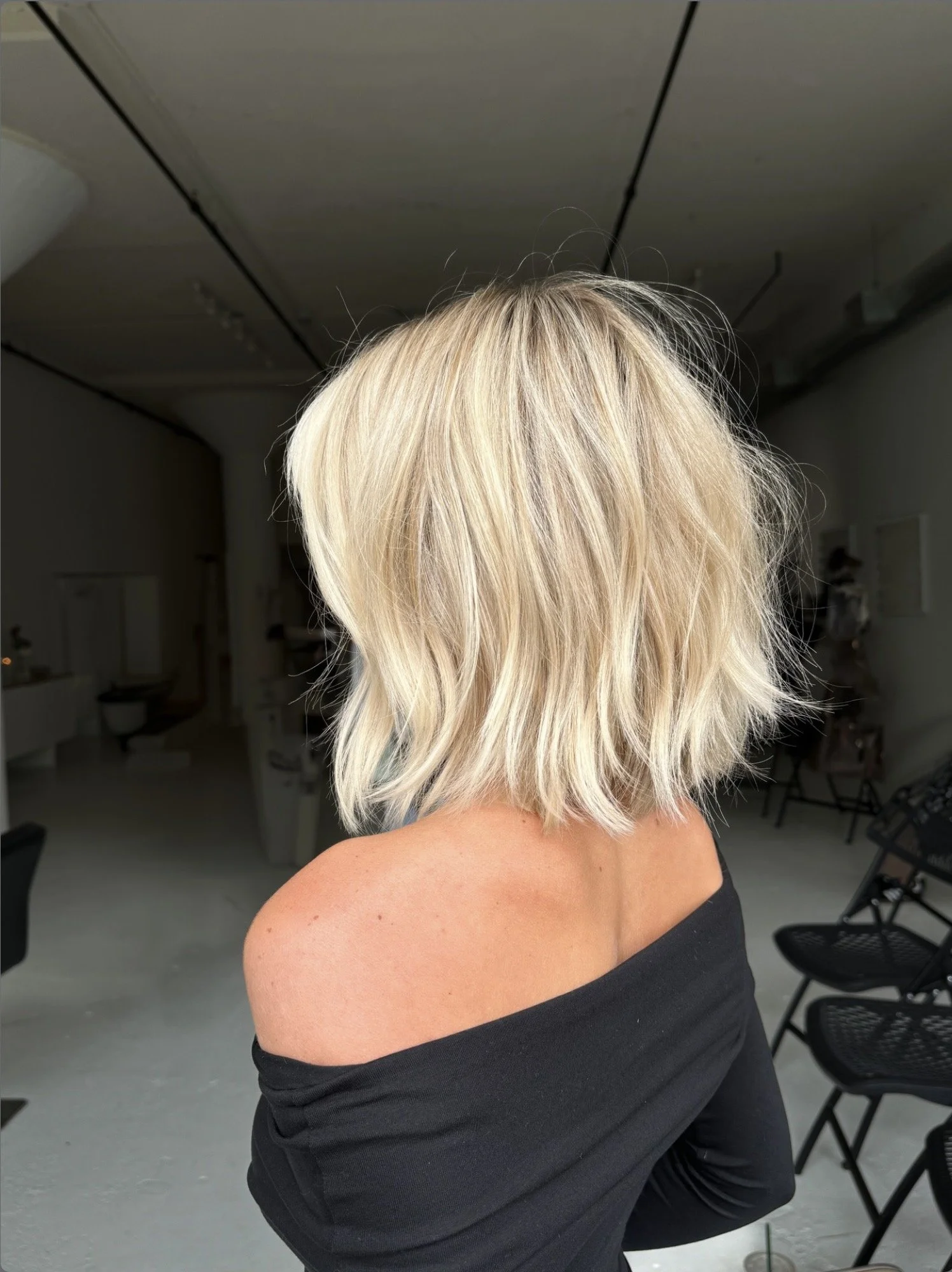 A person with short, messy blonde hair, wearing a black off-the-shoulder top, sitting in a room with chairs and a gray floor.