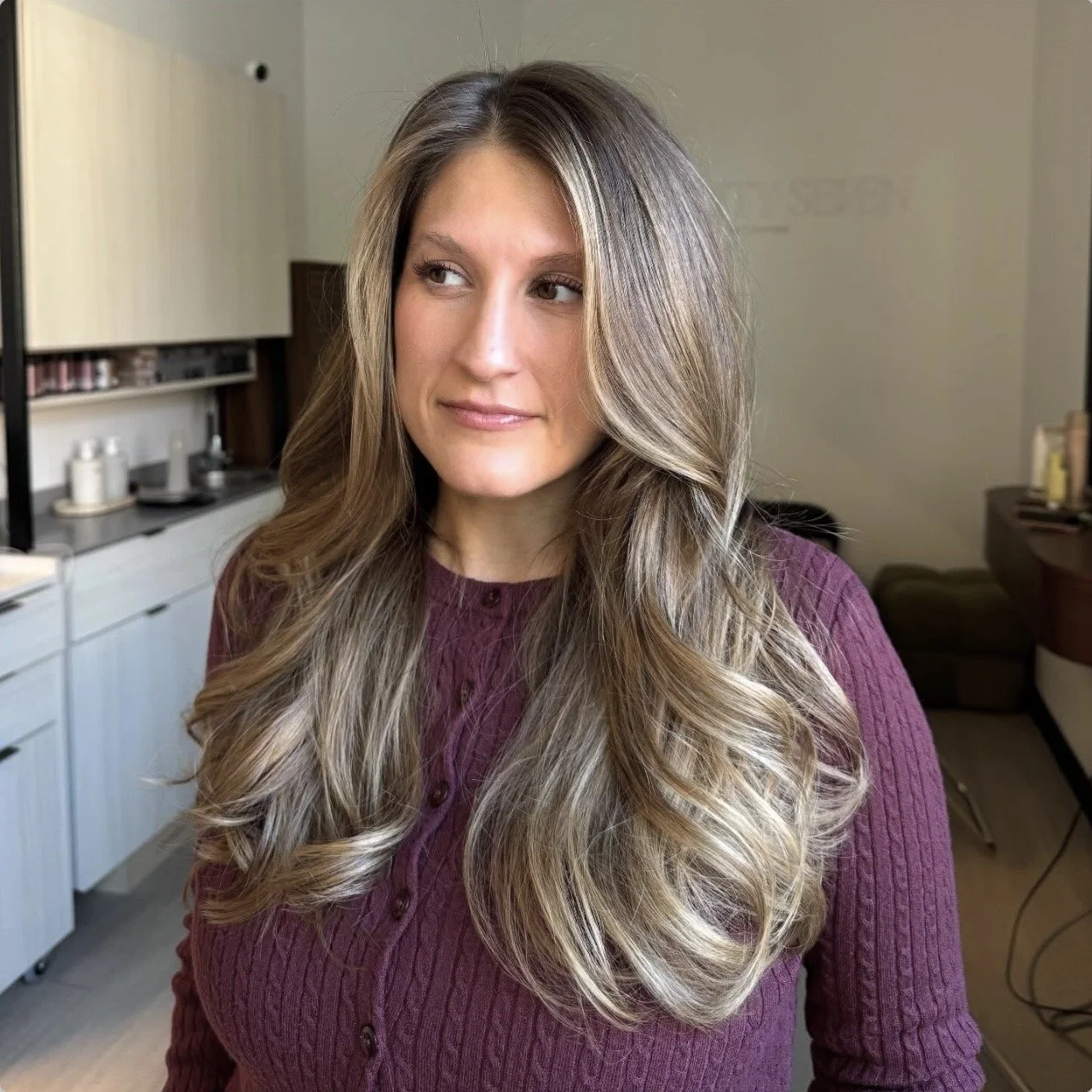 A woman with long wavy blonde hair wearing a purple sweater looks to the side in a room with kitchen and living room furniture.