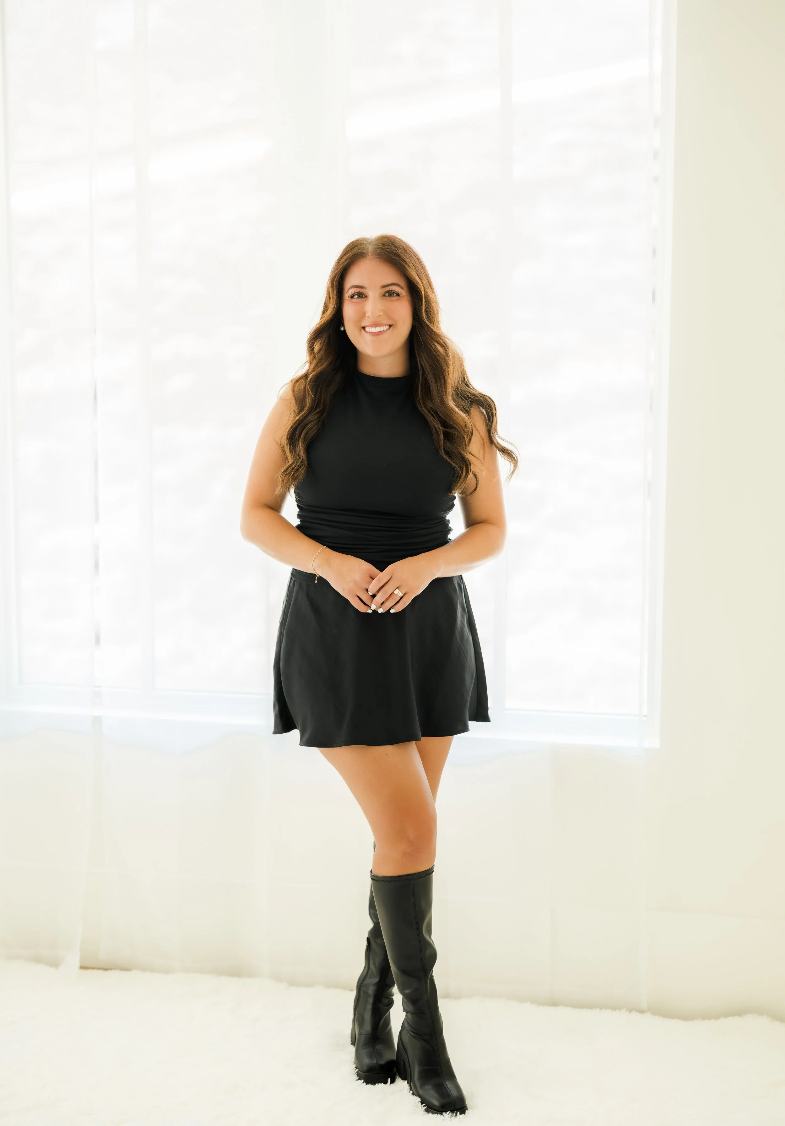 A woman with long wavy brown hair smiling, dressed in a black sleeveless top and a black skirt, wearing black knee-high boots, standing in front of white curtains with a bright window behind her.