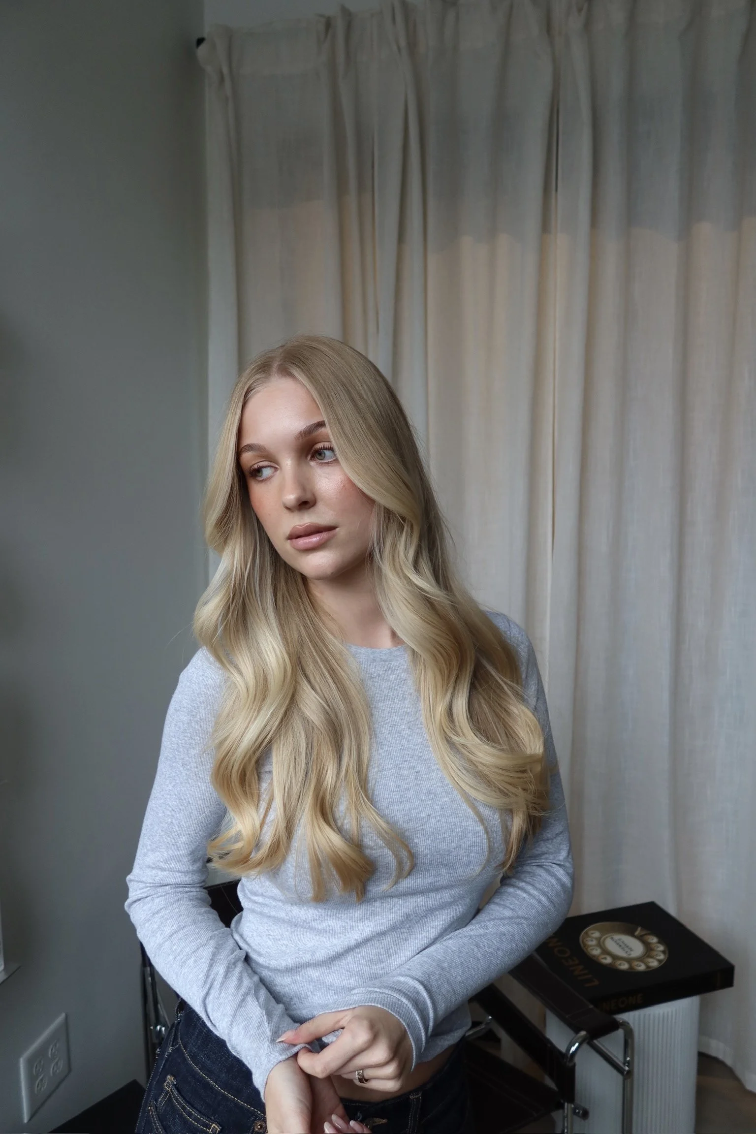 A young woman with long blonde wavy hair and light skin is looking slightly to her right. She is wearing a light gray long-sleeve top. Behind her is a cream-colored curtain and part of a room with gray walls. She is standing near a table with a black