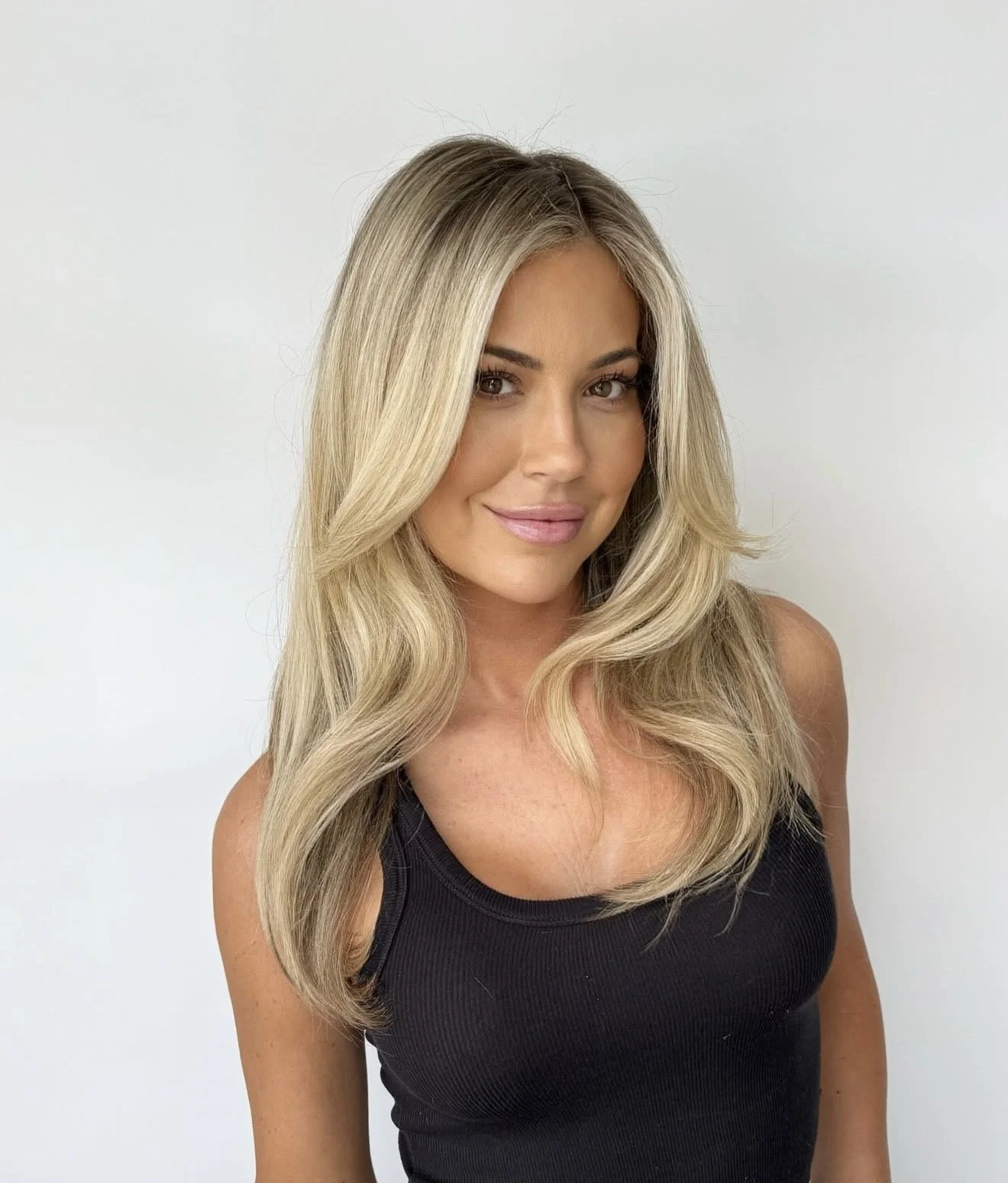 A woman with long, wavy blonde hair, wearing a black sleeveless top, standing against a plain white background, smiling softly.