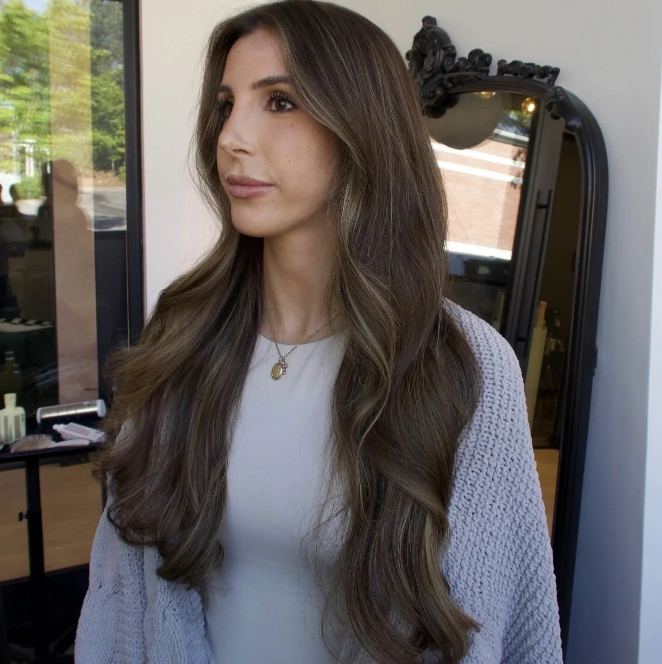 A woman with long, wavy brown hair posing in what appears to be a salon or studio, with a mirror and shelves with beauty products behind her.