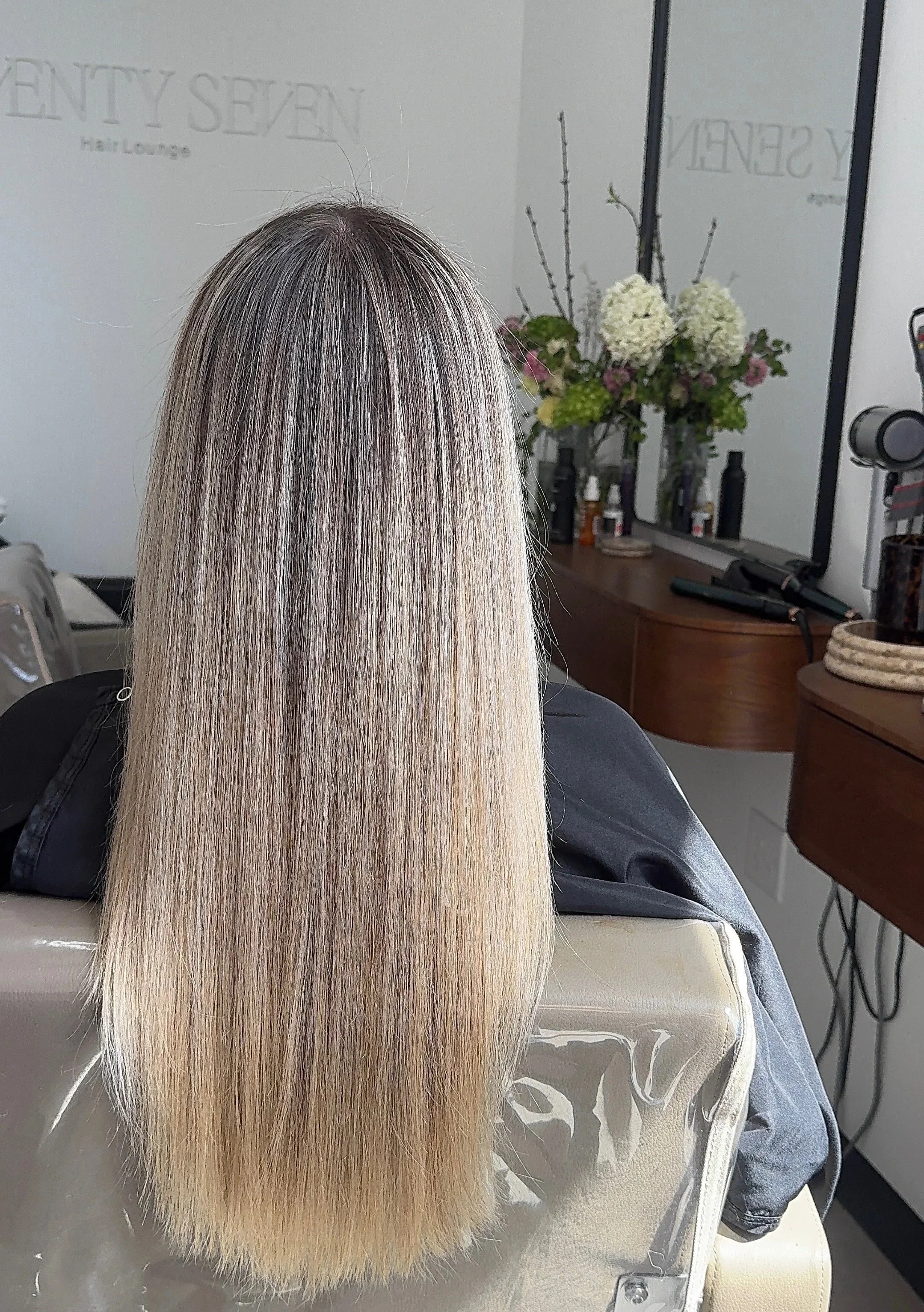 A person with long straight hair featuring a blend of black, gray, and blonde tones, sitting in a salon chair with a mirror and floral decoration in the background.