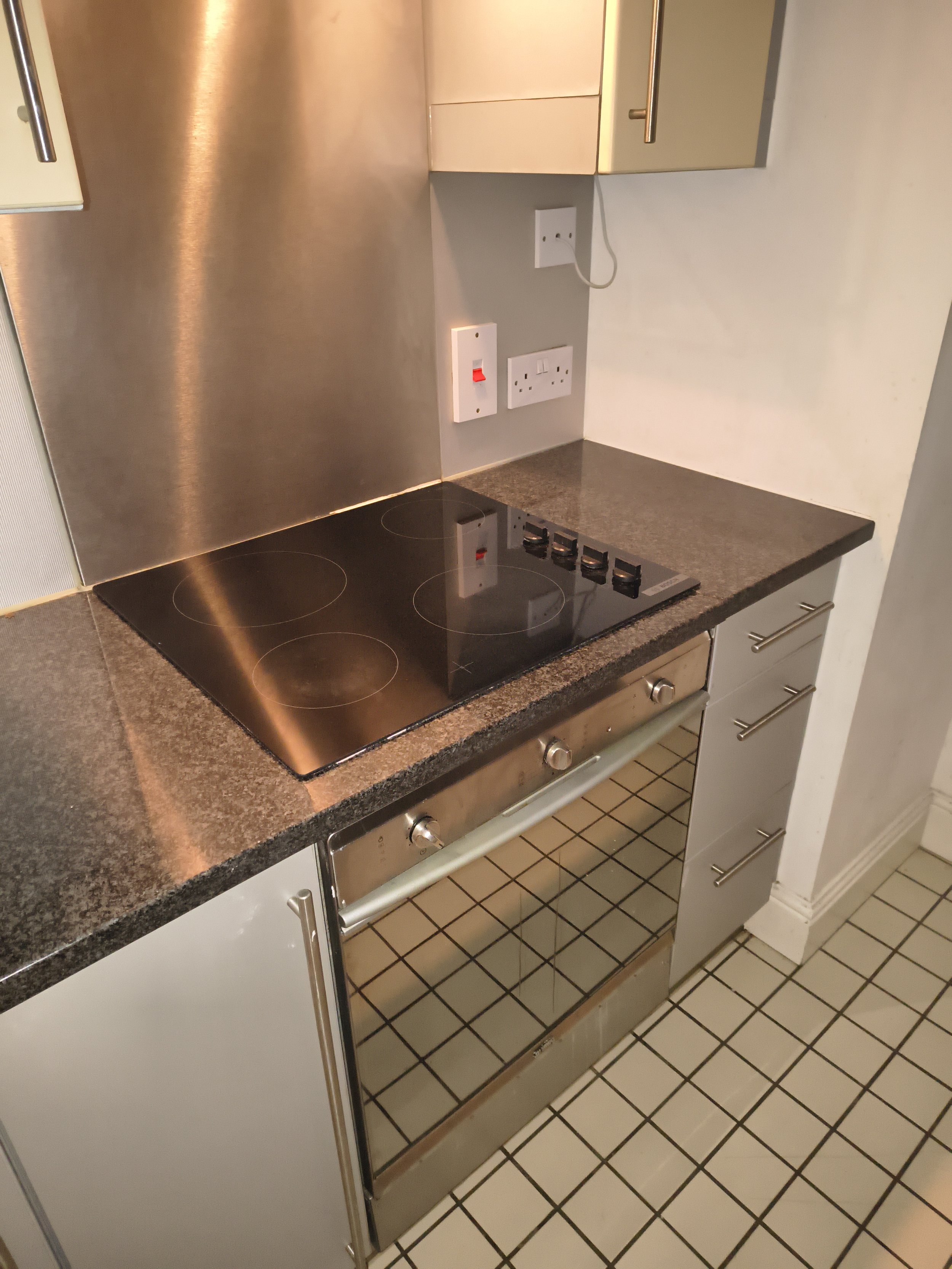 A kitchen stovetop with four electric burners, a stainless steel oven, gray cabinets, a brown speckled countertop, and tiled floor with a white and black pattern.