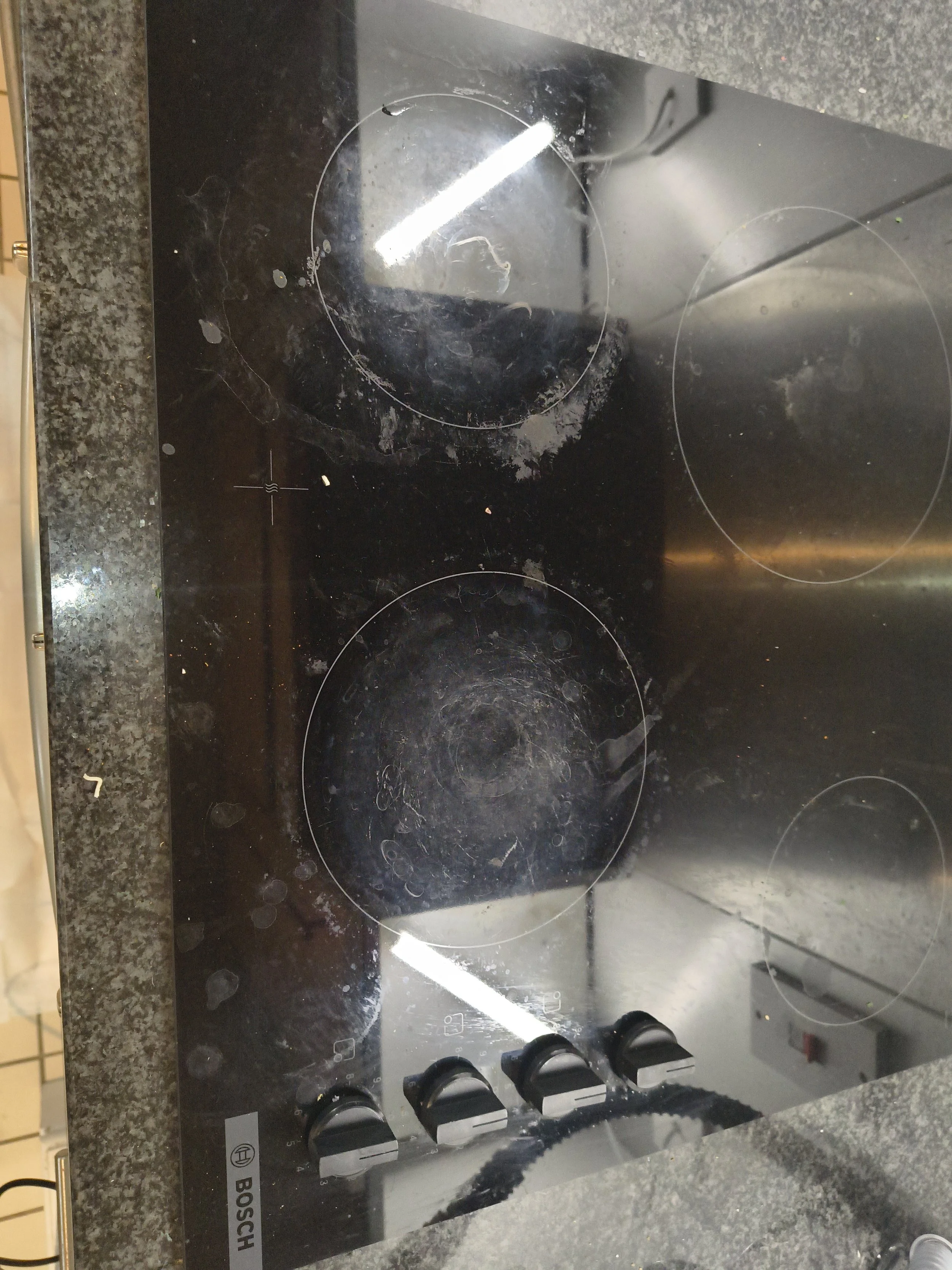Black glass electric stovetop with four burners and four control knobs, some burners have visible cleaning marks and residue.