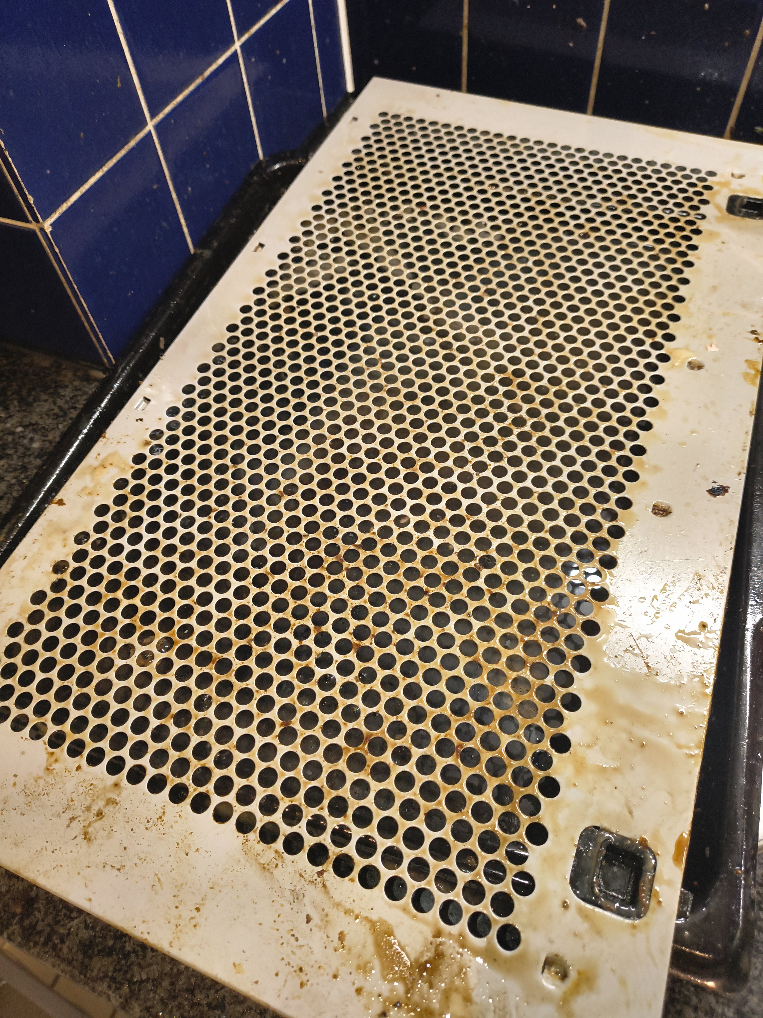 Dirty and greasy stovetop cover with small round holes, showing stains and rust around the edges.