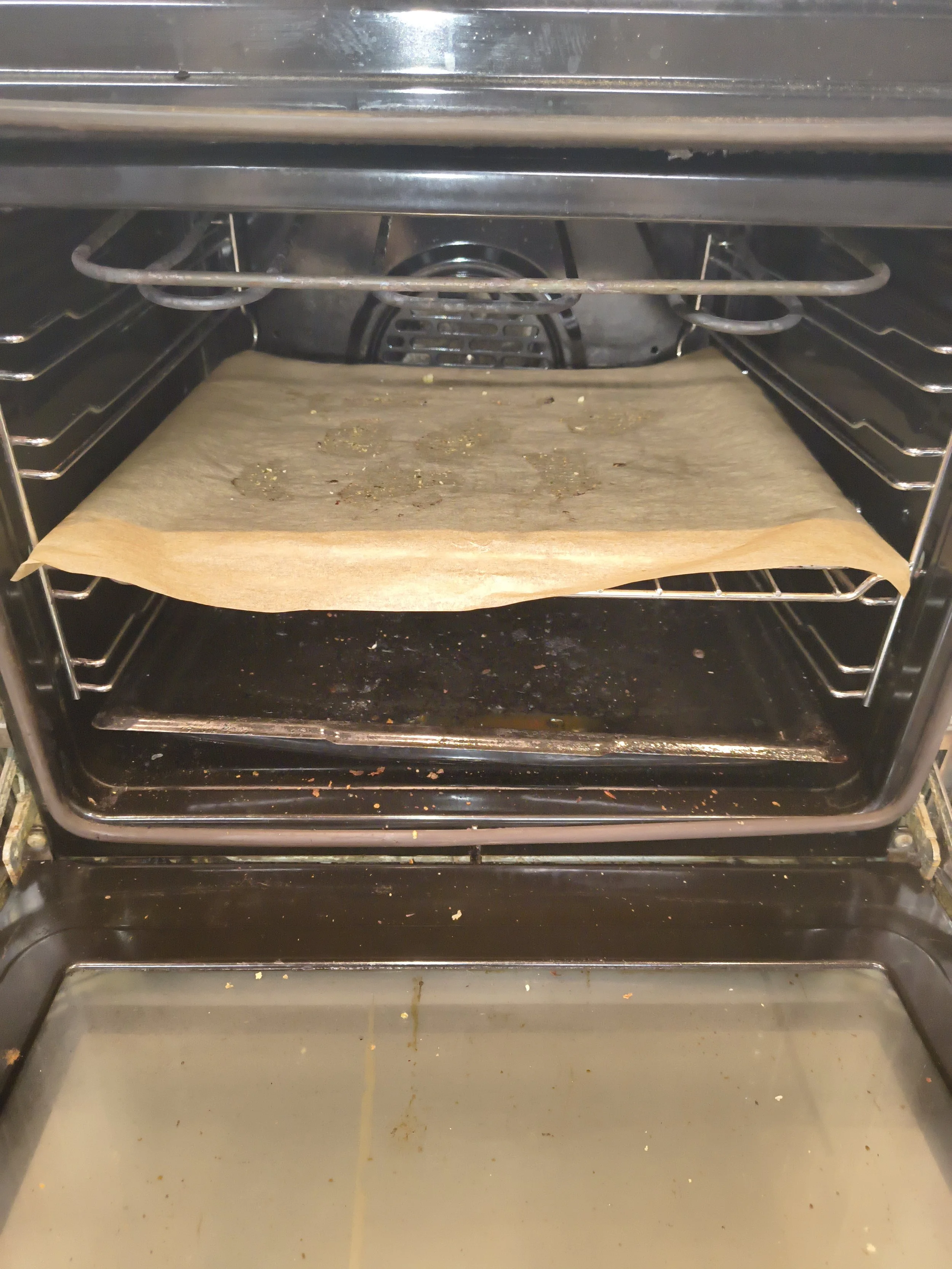 An empty oven with a baking sheet on the lower rack, a piece of parchment paper on the middle rack, and some burnt residues inside.