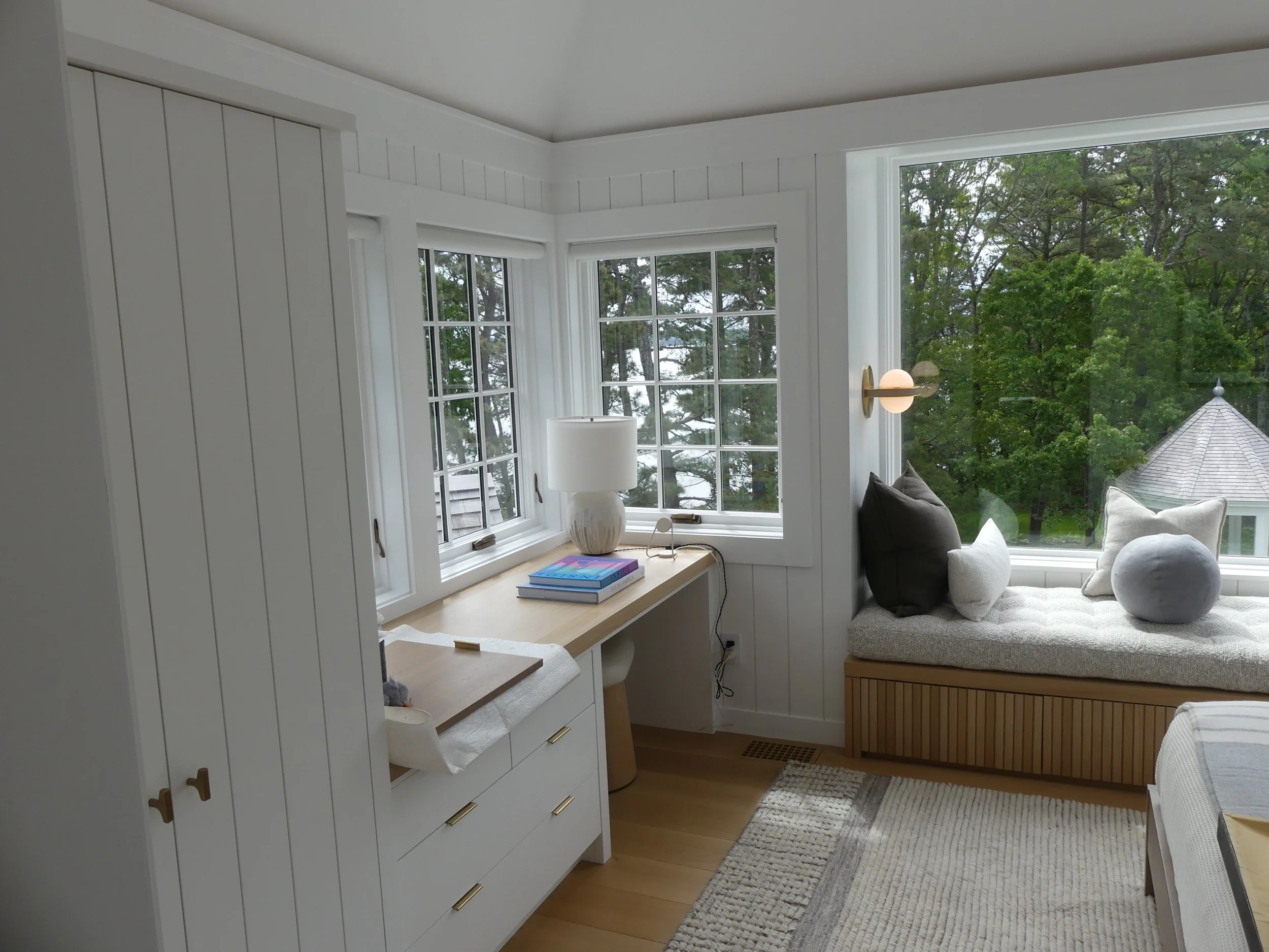 A cozy window nook with a cushioned bench and pillows, surrounded by large windows with a view of trees and a building with a conical roof outside.