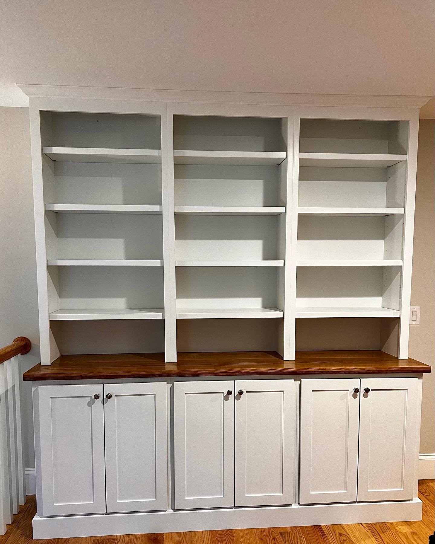 White wooden bookshelf with three open shelves at the top and enclosed cabinets at the bottom, topped with a wooden countertop, against a beige wall.