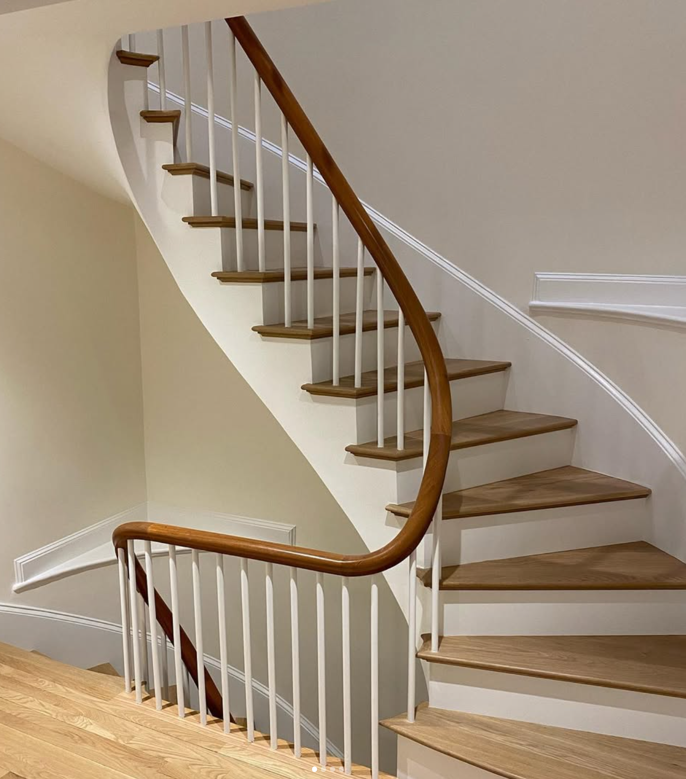 Indoor staircase with wooden steps, white risers, and a curved wooden handrail on white spindles against beige walls.