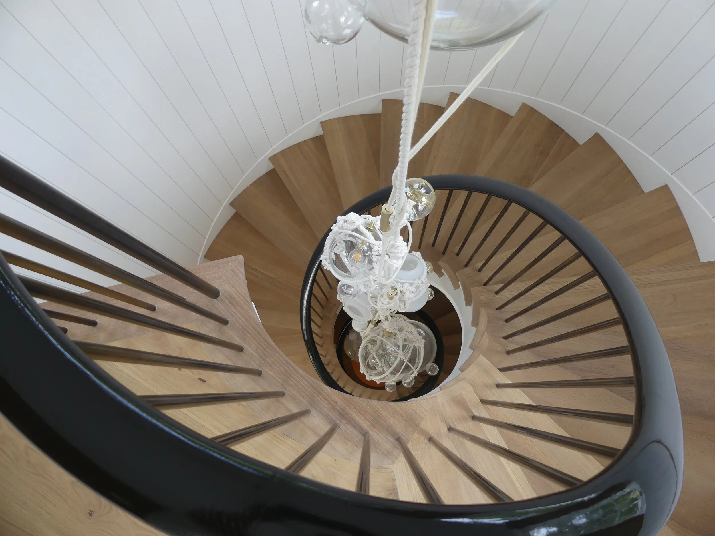 View looking down a spiral staircase, showing the wooden steps, black handrail, and a white decorative chandelier hanging at the center.