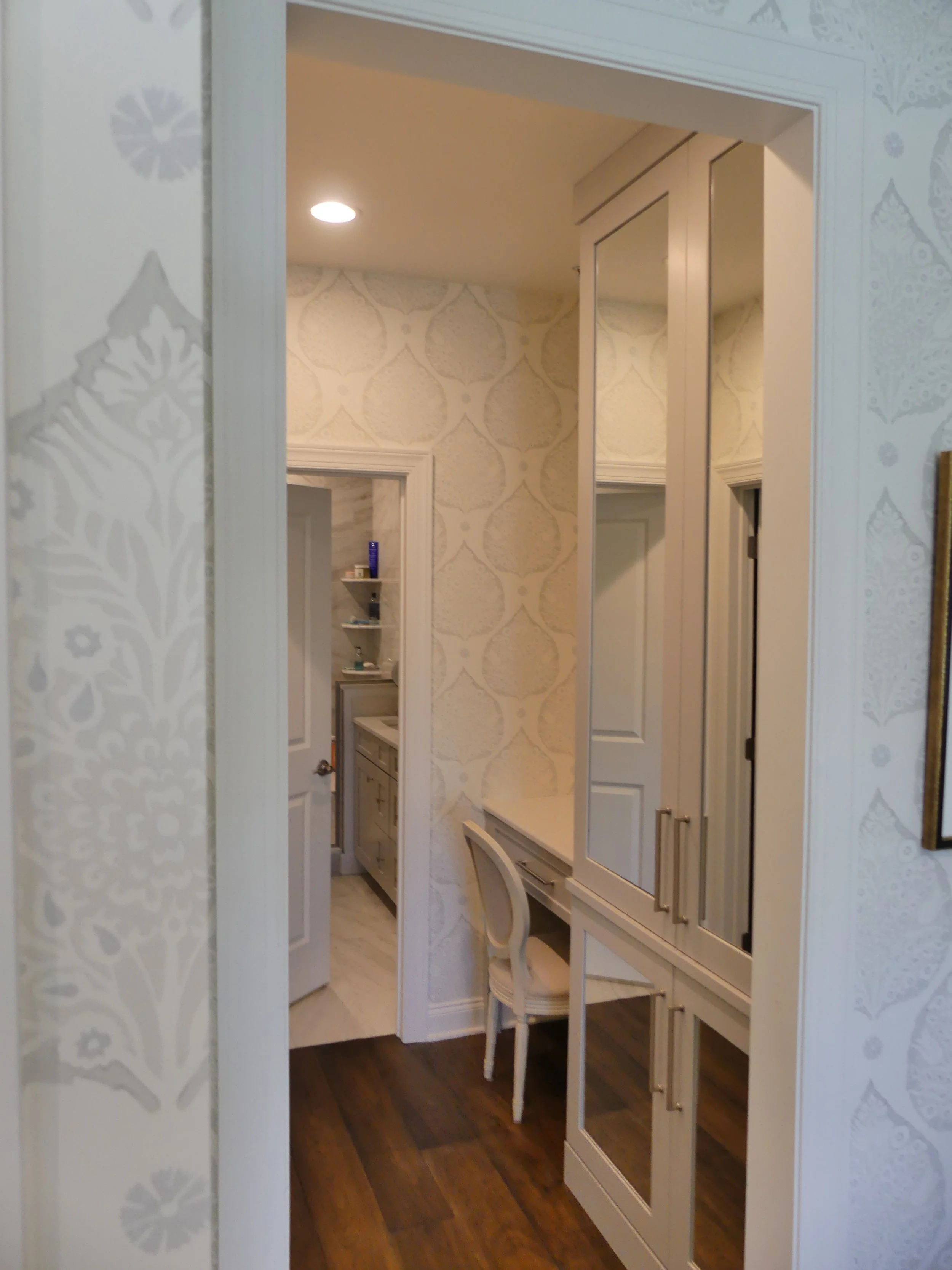 View through an open doorway into a room with a built-in wardrobe, a small vanity with a chair, patterned wallpaper, and a kitchen area visible in the background.