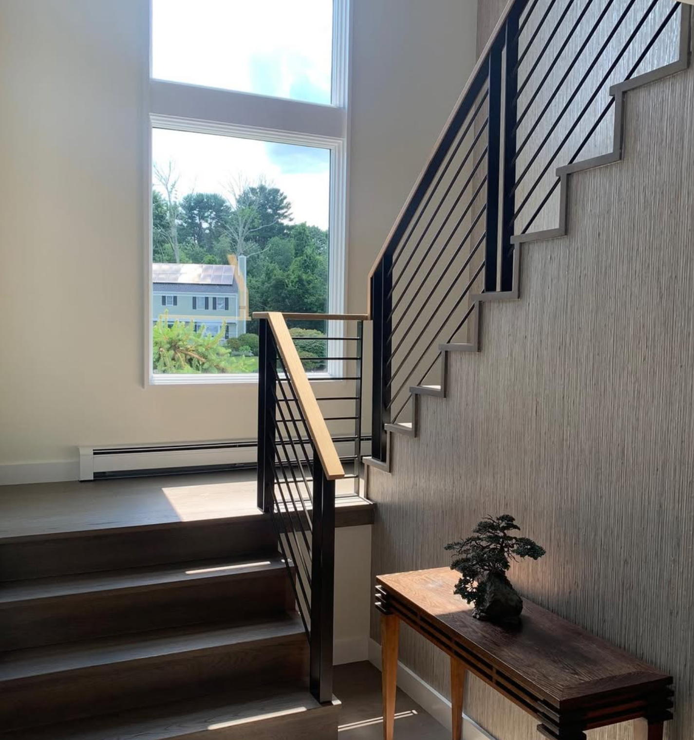 Interior view of a stairway with wooden steps, black metal railings, large windows letting in sunlight, and a decorative bonsai tree on a wooden table.