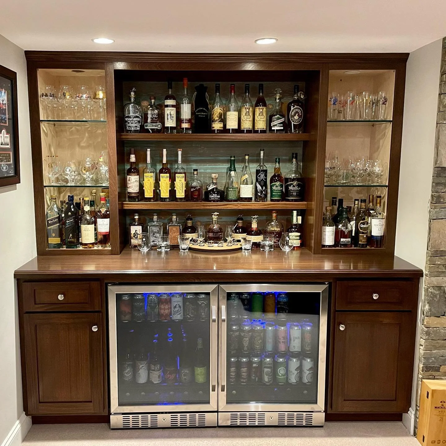 a home bar with a wooden cabinet, glass doors, and shelves filled with liquor bottles and glassware, with a mini fridge below containing beverages