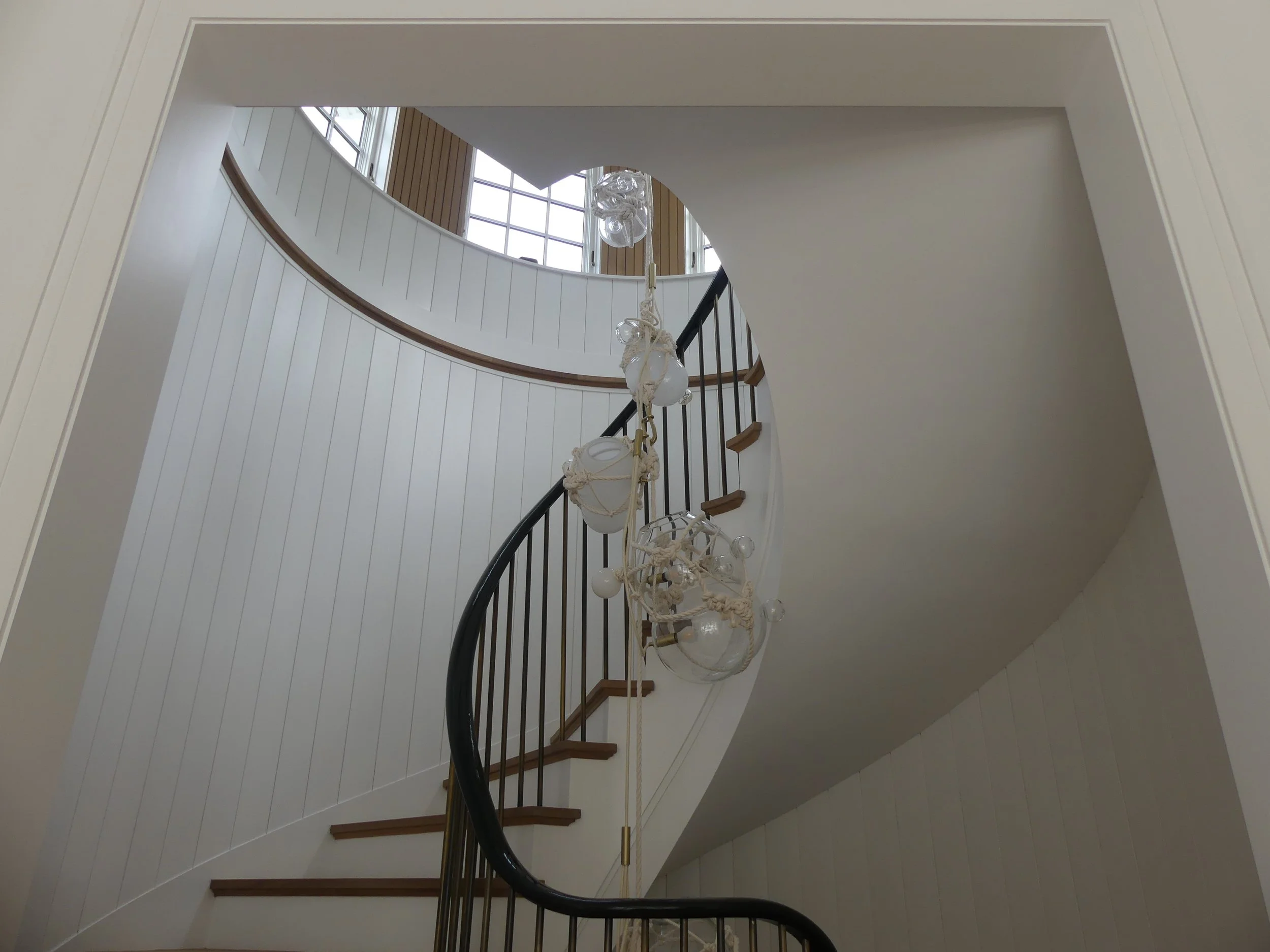A spiral staircase with wooden steps and black railing, viewed from the bottom, with a modern chandelier hanging in the center and large windows at the top letting in natural light.