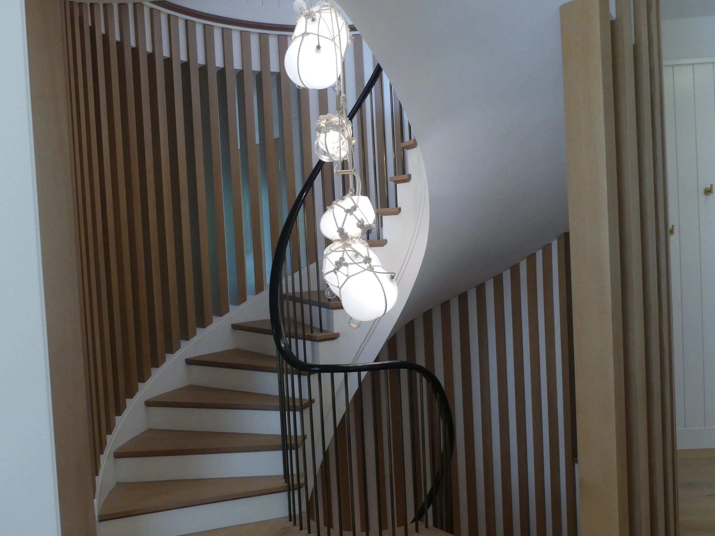 Interior view of a curved staircase with wooden steps, black railing, wooden vertical slats on the wall, and a modern chandelier with multiple white globes hanging from the ceiling.