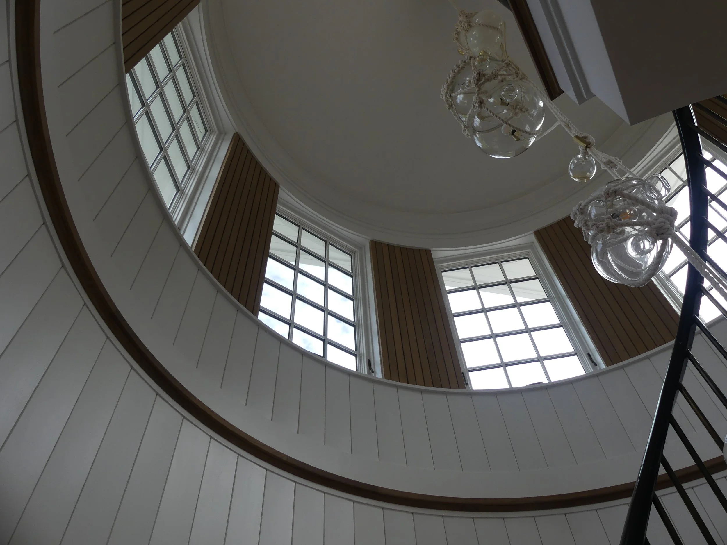 Looking up at a spiraling staircase with white railing, wooden accents, large windows, and a decorative chandelier hanging from the ceiling.