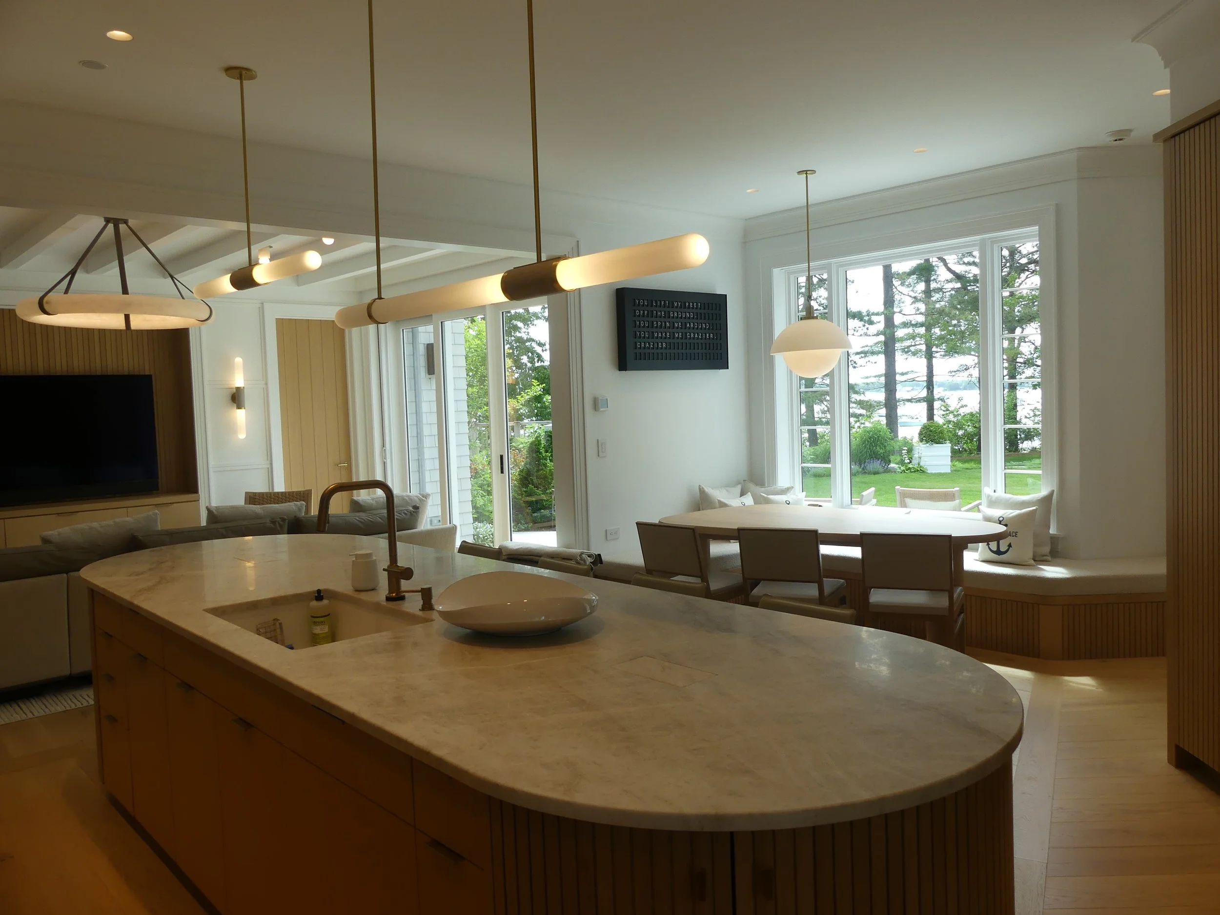 Open-concept kitchen and dining area with large windows showing greenery outside, a marble island with a faucet and bowl, modern lighting fixtures, and a television on the wall.