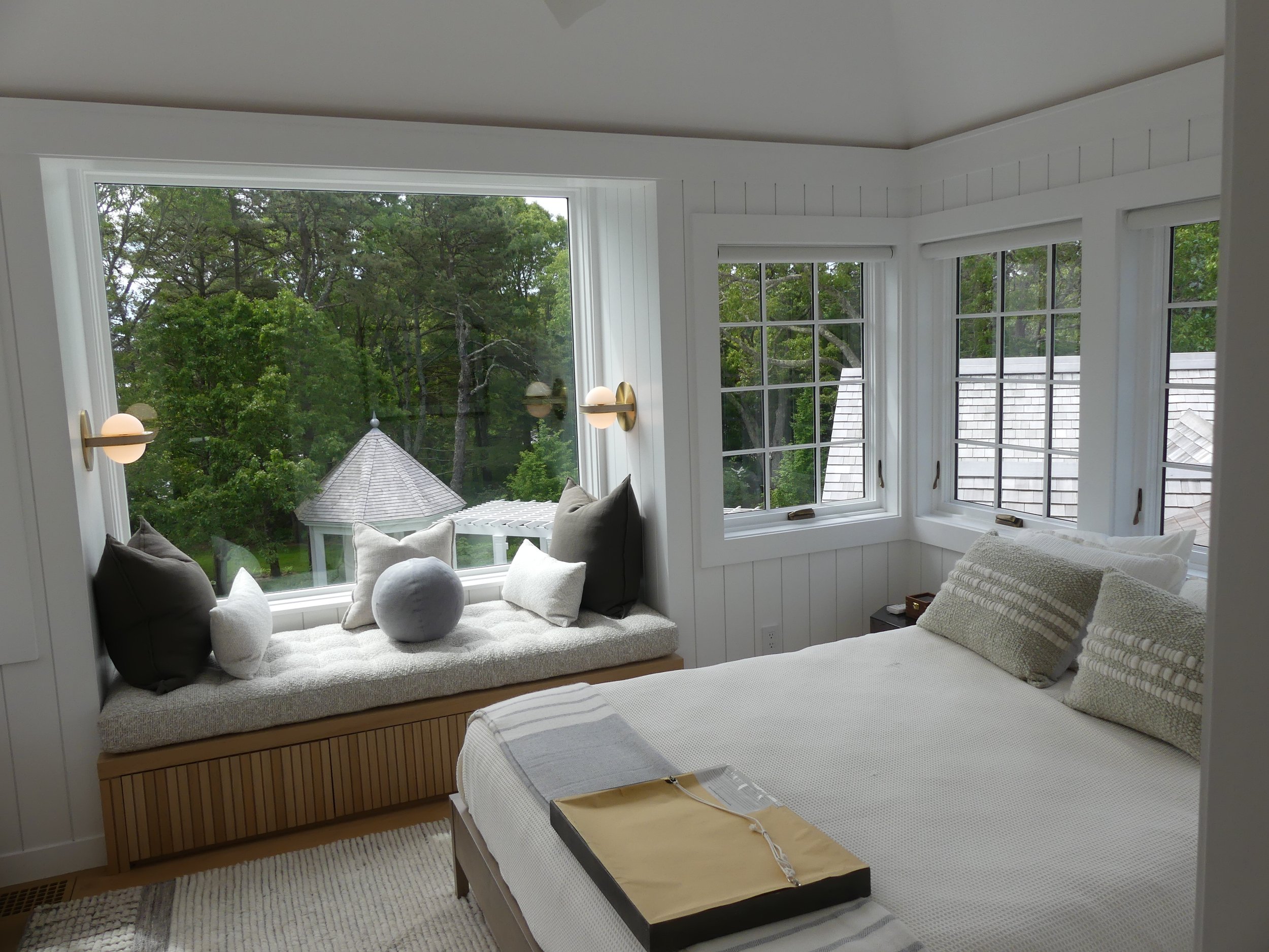 A cozy bedroom with a window seat with pillows and a view of a backyard with trees and a gazebo, and a bed with pillows and a tray on top.